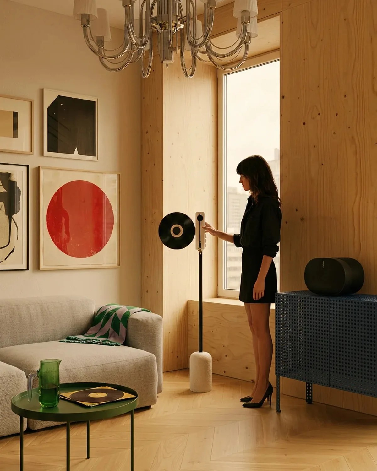 Woman in black dress operating a modern vertical record player in a wood-paneled living room with beige sofa and green side table.