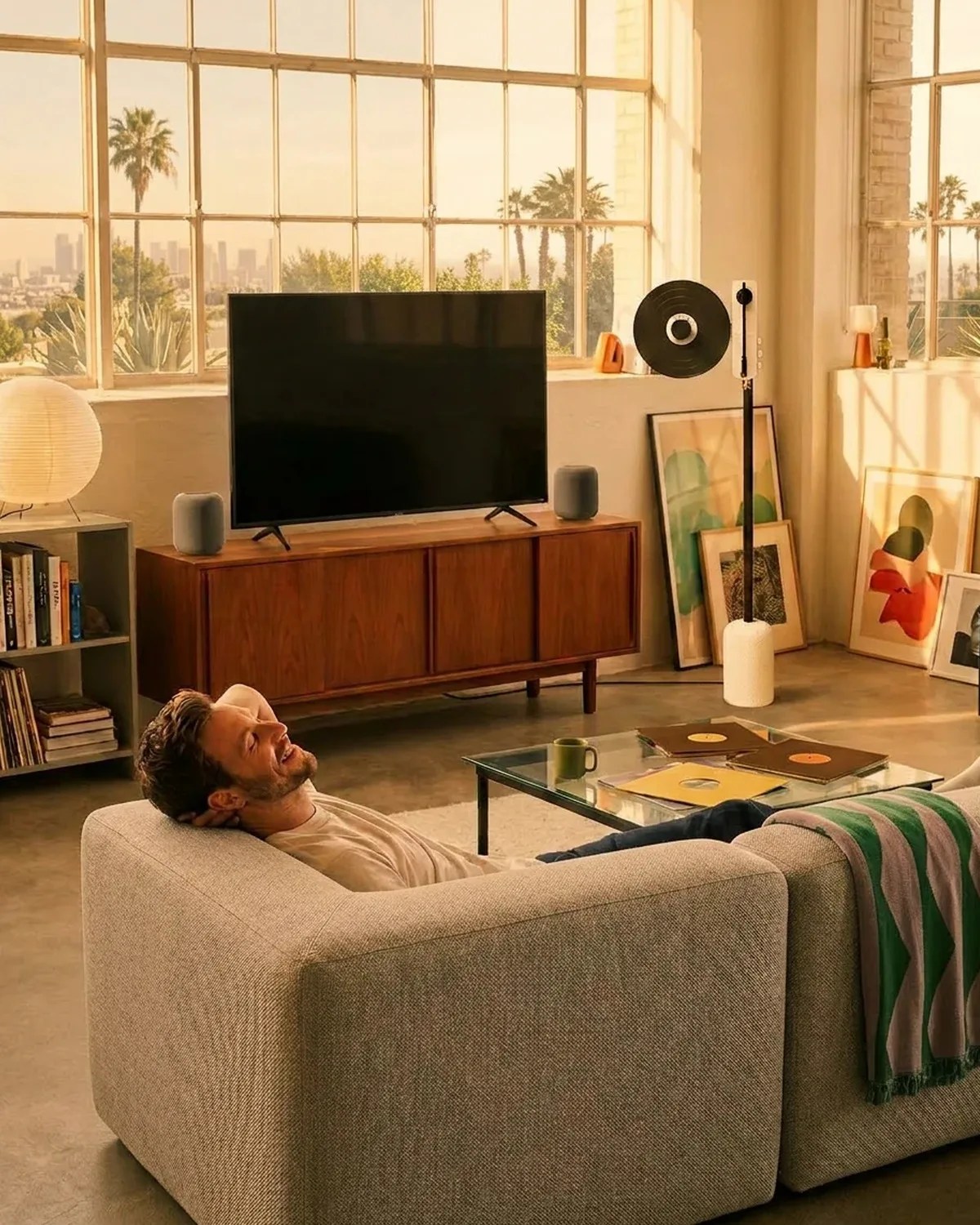 Man reclining on a beige fabric sofa with a green and gray striped blanket in a sunlit living room with a TV, wooden cabinet, and vinyl records.