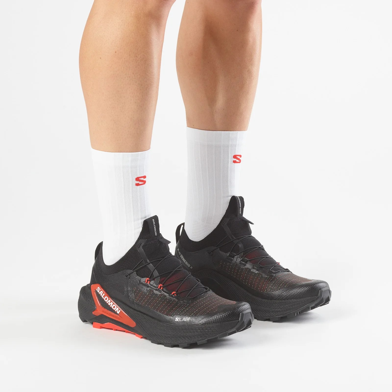Black Salomon trail running shoes with red accents worn with white crew socks on legs.