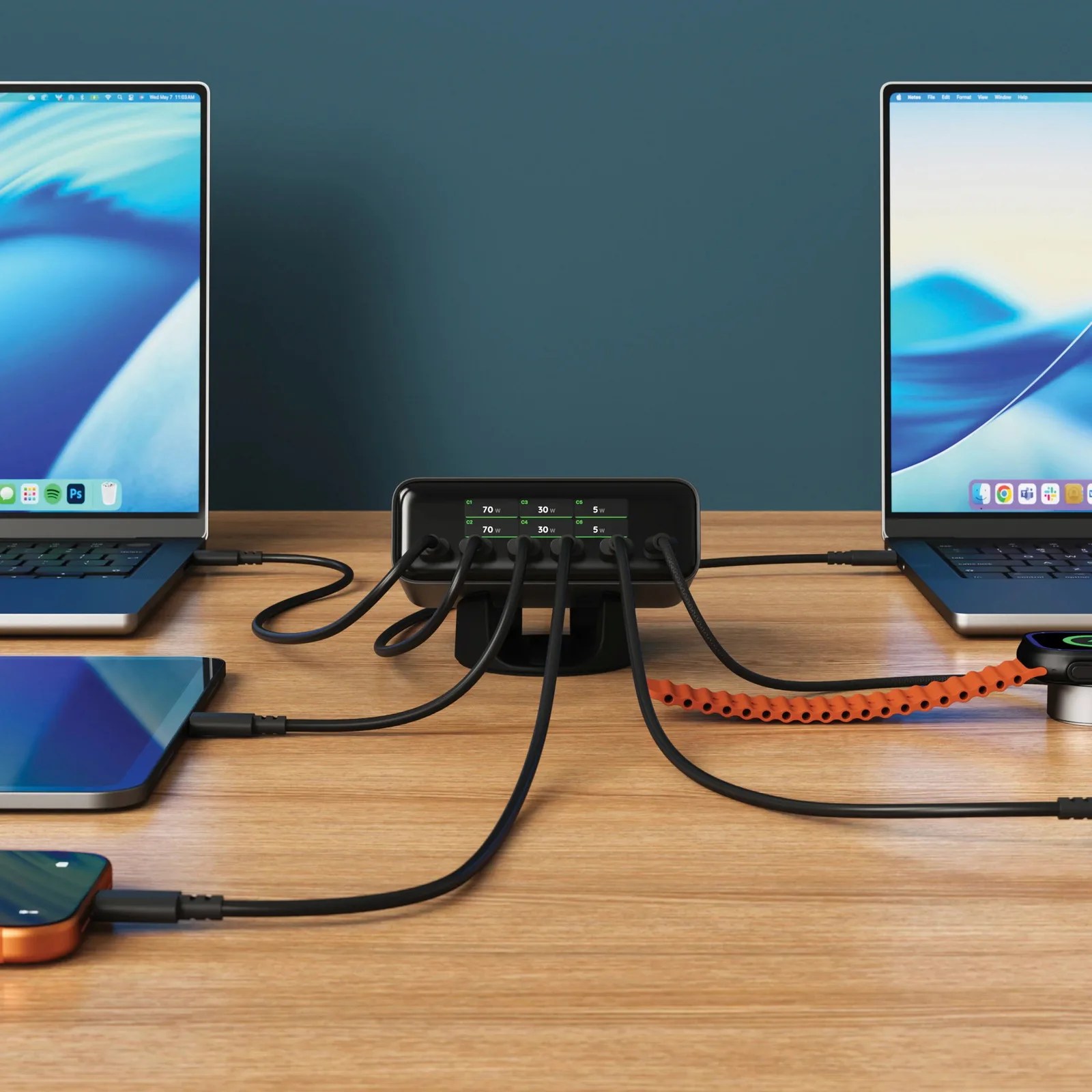 Black multi-port charging hub with digital power display connected to laptops, smartphone, and smartwatch on a wooden desk.
