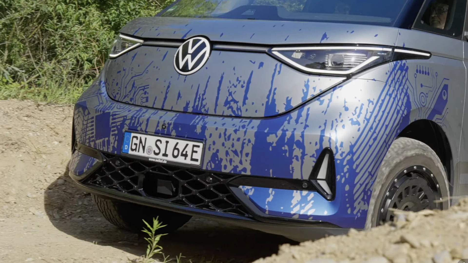 Front view of a silver Volkswagen electric vehicle with blue circuit-like patterns and a German license plate on a dirt road.