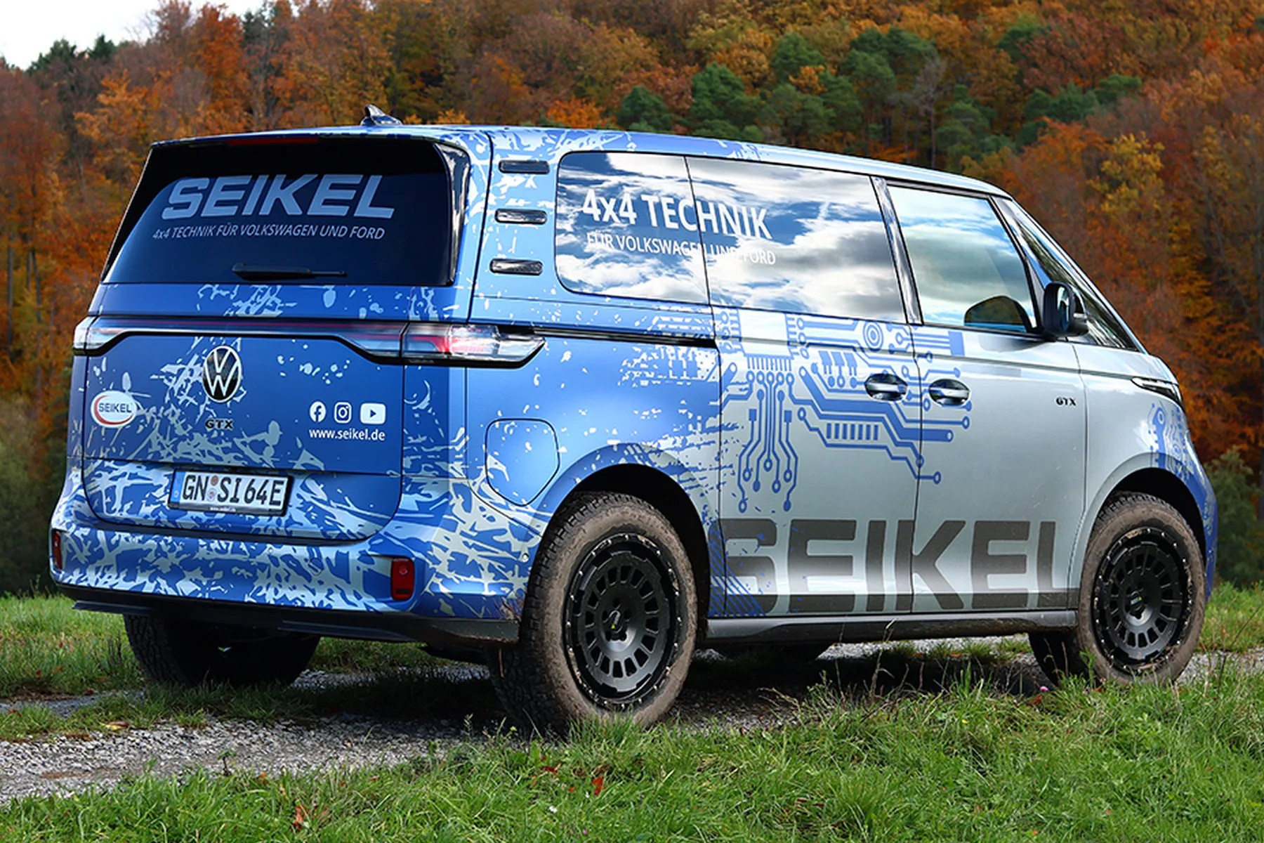 Blue and silver Volkswagen van with Seikel branding and circuit board graphics parked on grass with autumn trees in background.