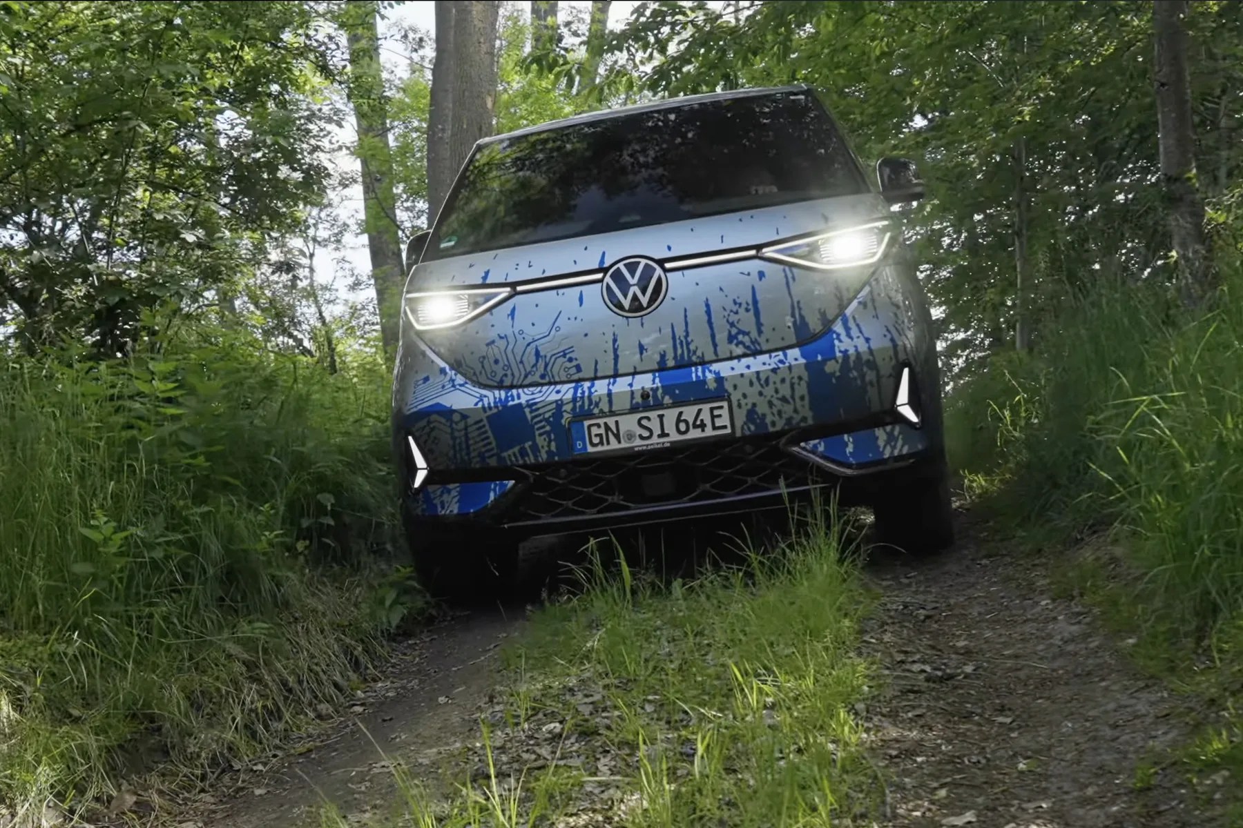 Front view of a blue Volkswagen electric van with headlights on, driving on a narrow forest trail.