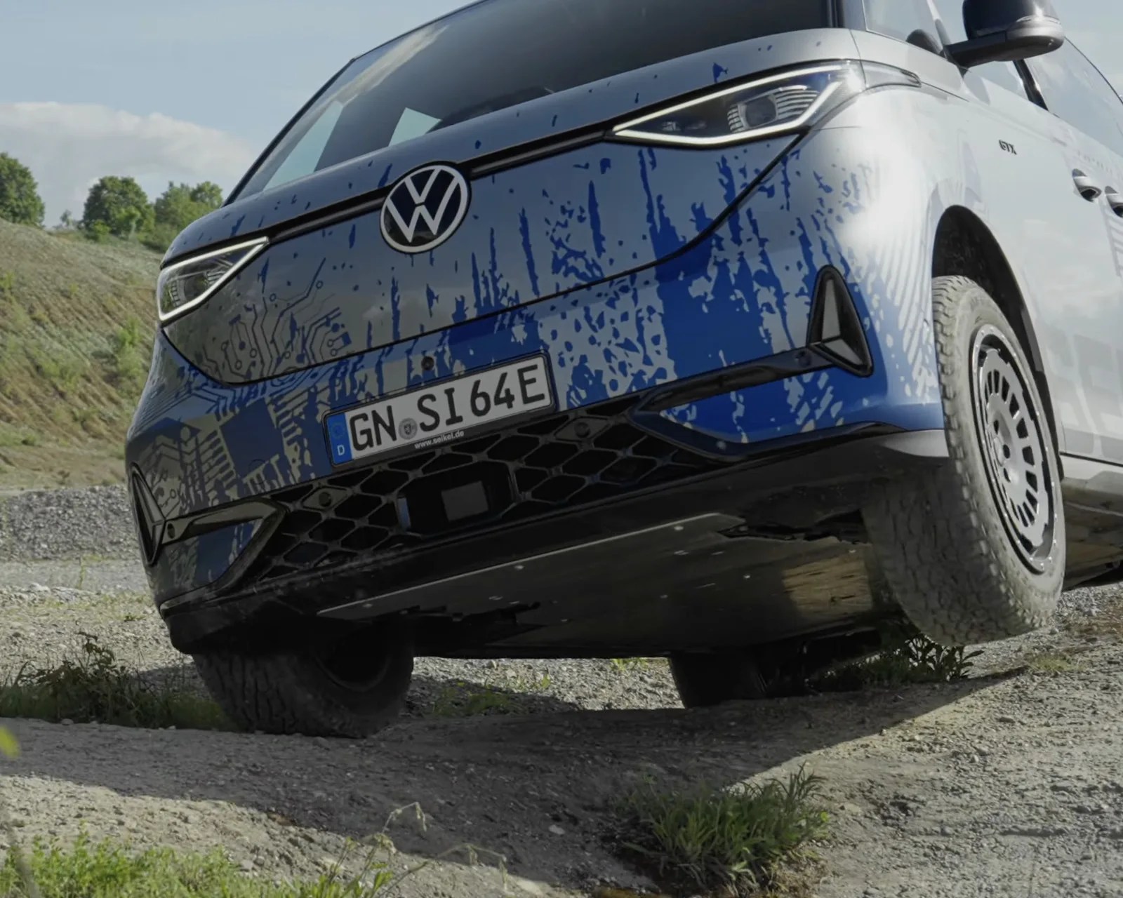Front view of a blue Volkswagen electric vehicle with circuit-like patterns on the hood, driving on a gravel path.
