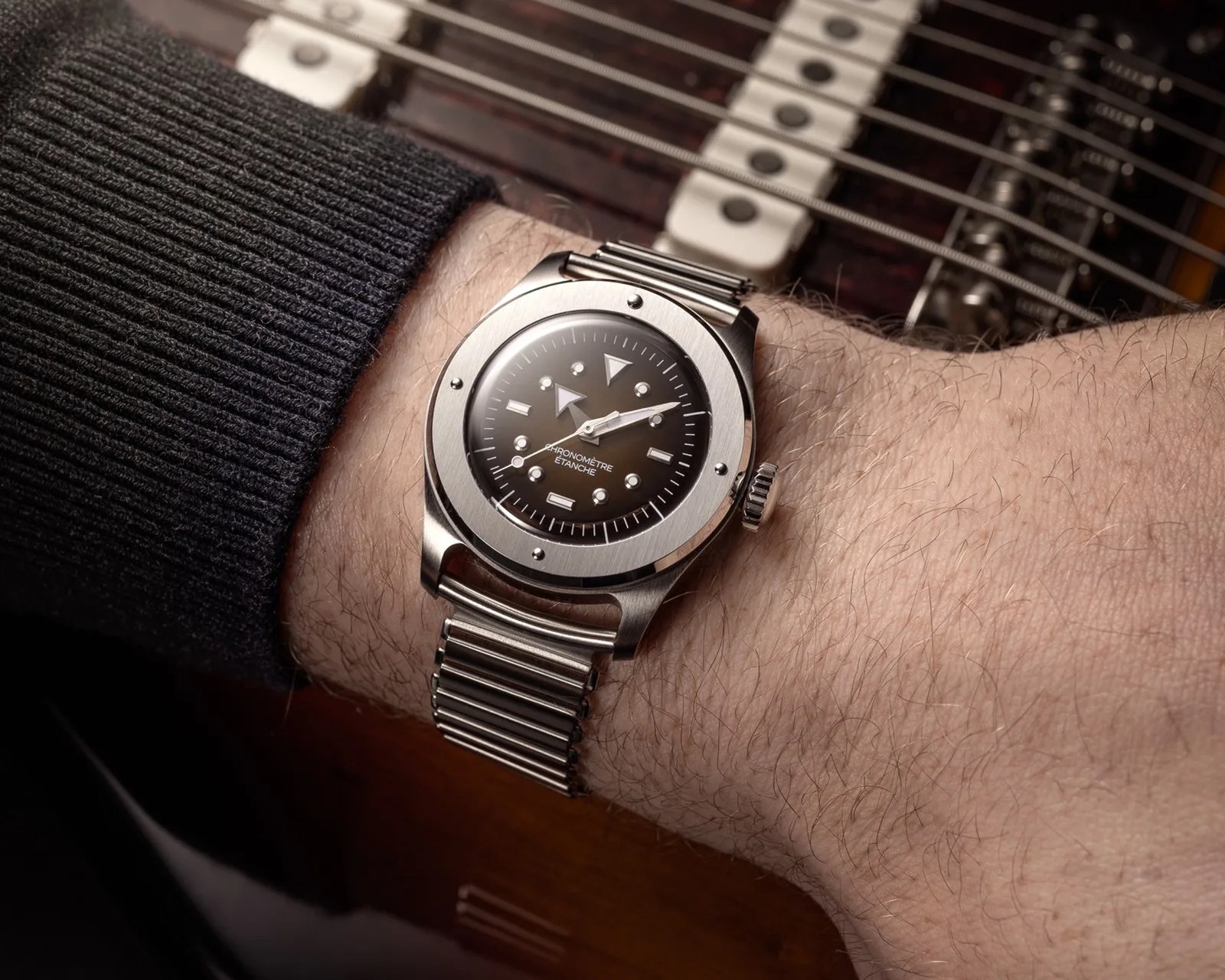 Silver wristwatch with black dial and metal expandable band worn on a wrist over a guitar background.