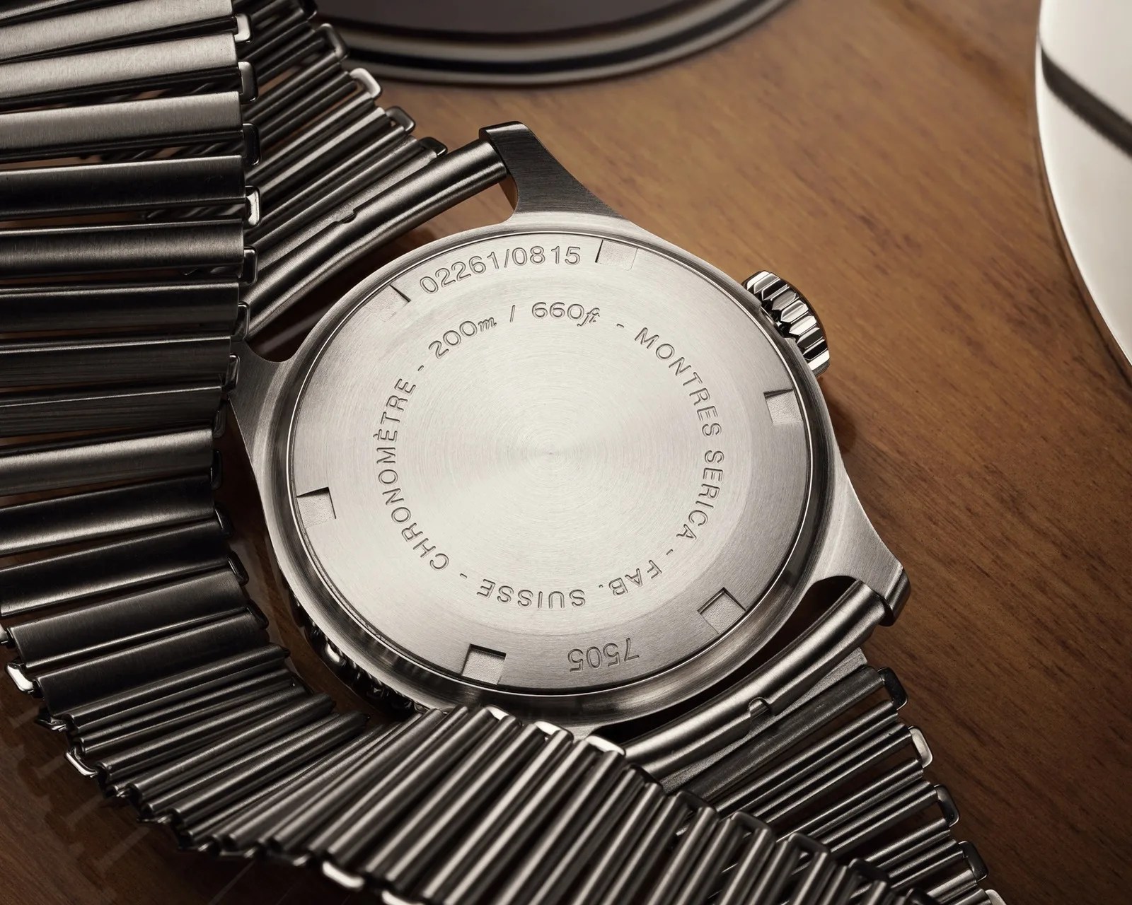 Brushed stainless steel watch back with engraved text and a metal expansion bracelet on a wooden surface.