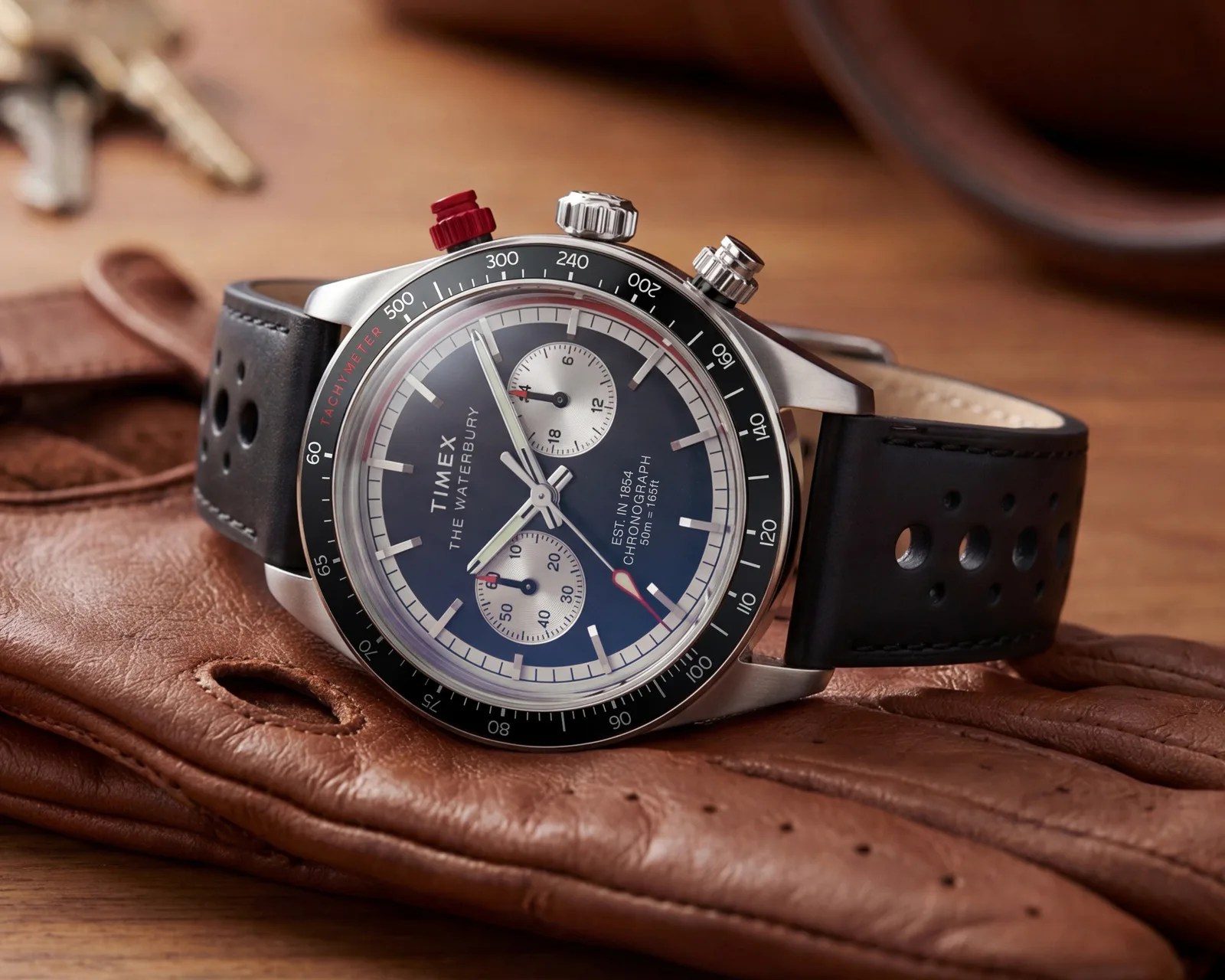 Timex chronograph watch with black perforated leather strap and black tachymeter bezel on brown leather gloves.