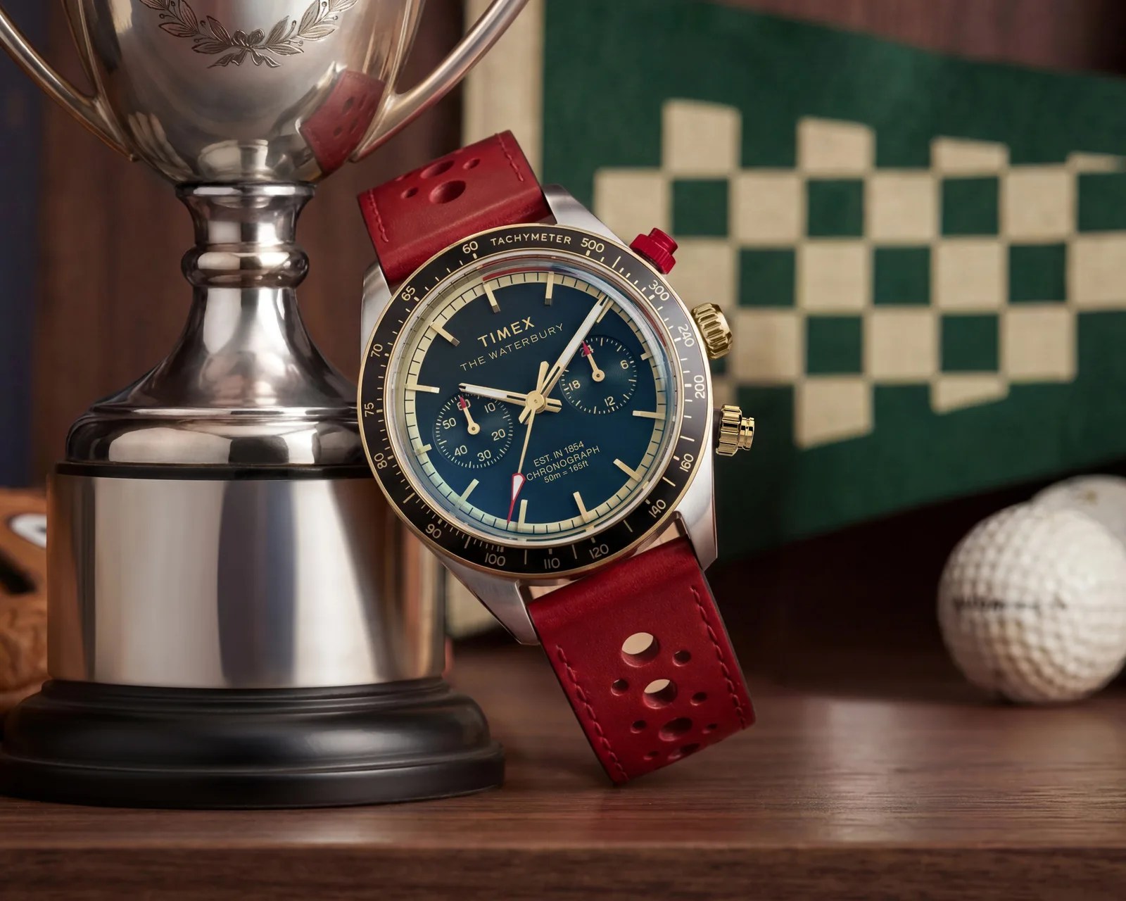 Chronograph Timex Waterbury watch with blue dial, gold accents, black tachymeter bezel, and red perforated leather strap leaning on a silver trophy.