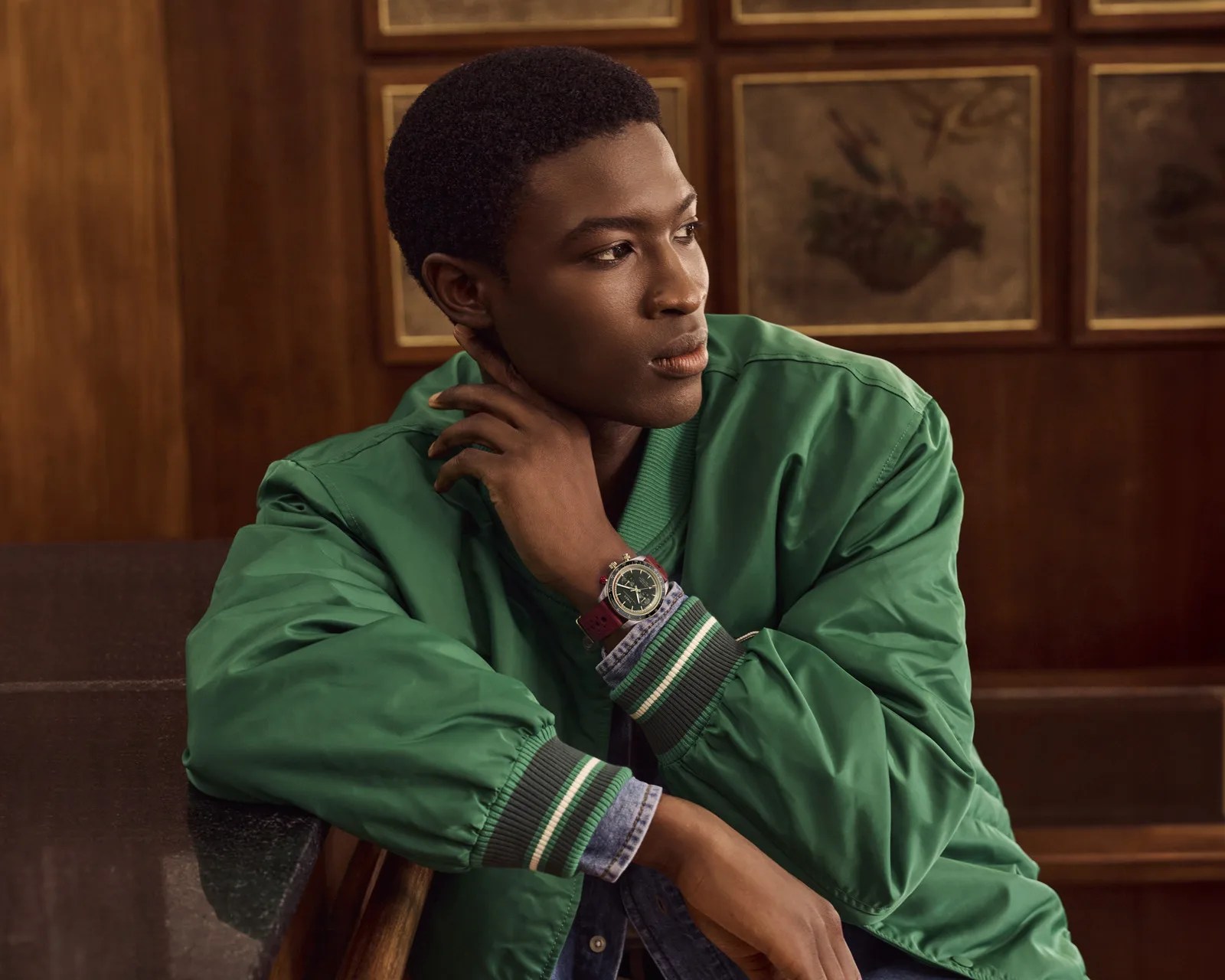 Person wearing a green jacket and denim shirt, resting arm on a table, with a wristwatch featuring a red strap.