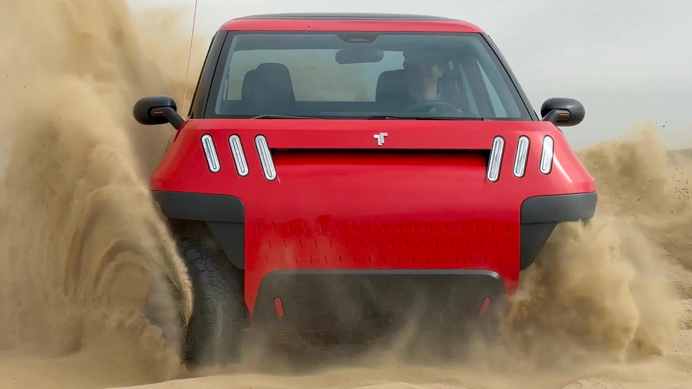 Red off-road vehicle kicking up sand with a boxy front and six vertical LED lights.