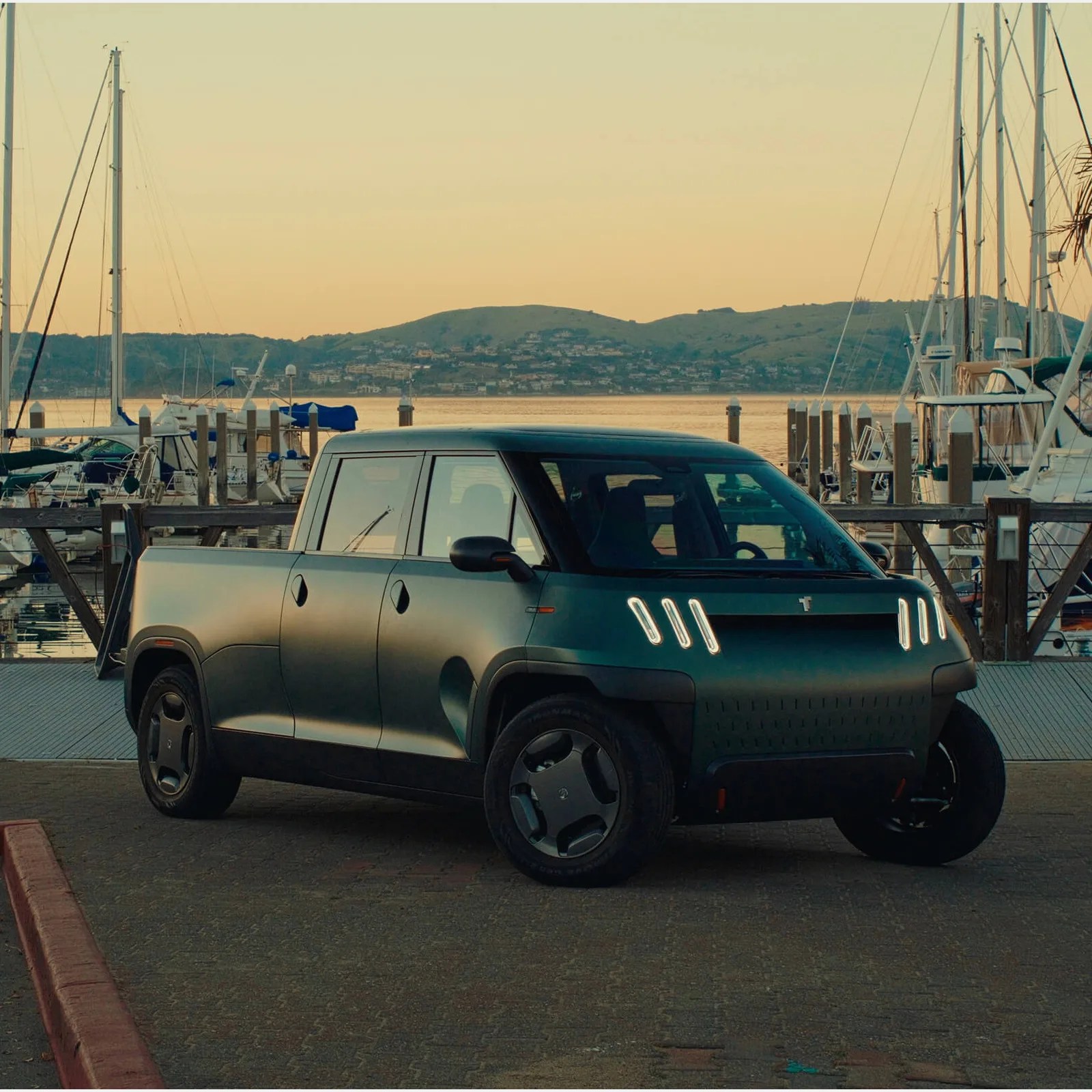 Matte dark green electric pickup truck with unique vertical LED headlights parked near a marina at sunset.