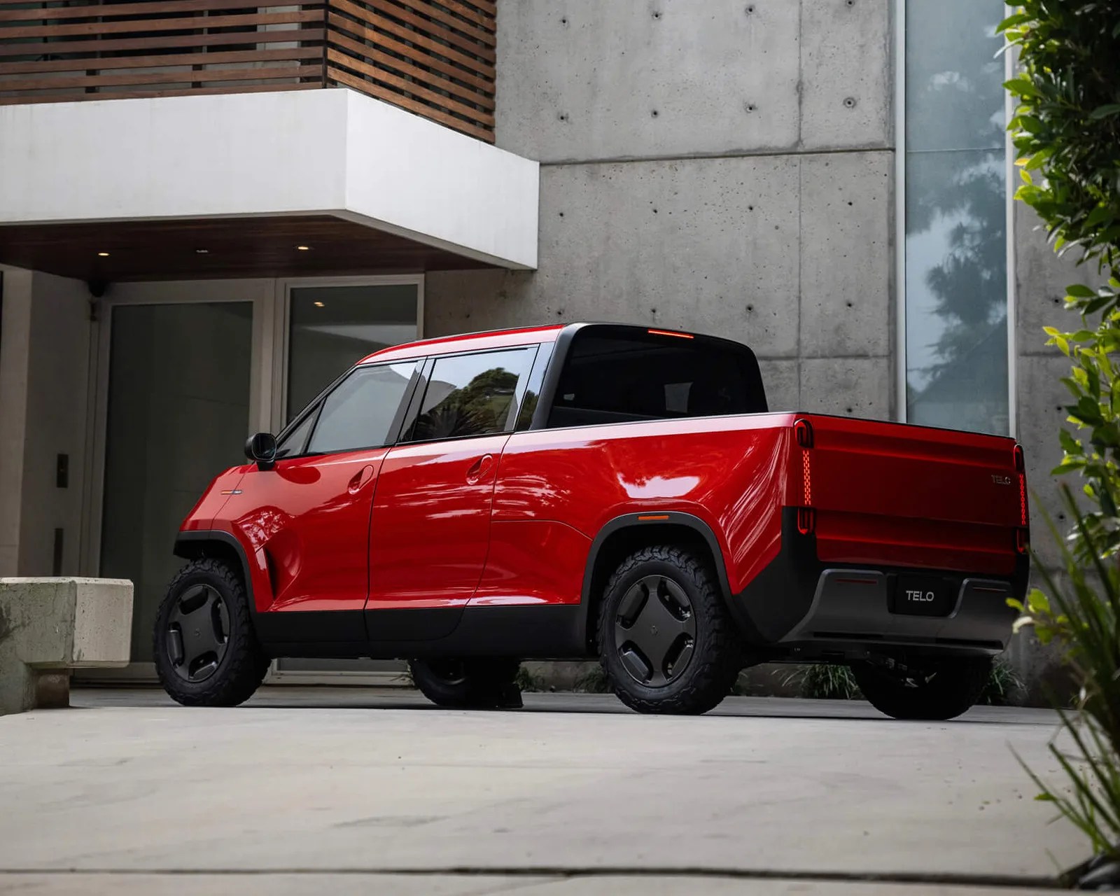 Red electric pickup truck with black roof and rugged tires parked in front of a modern concrete building.