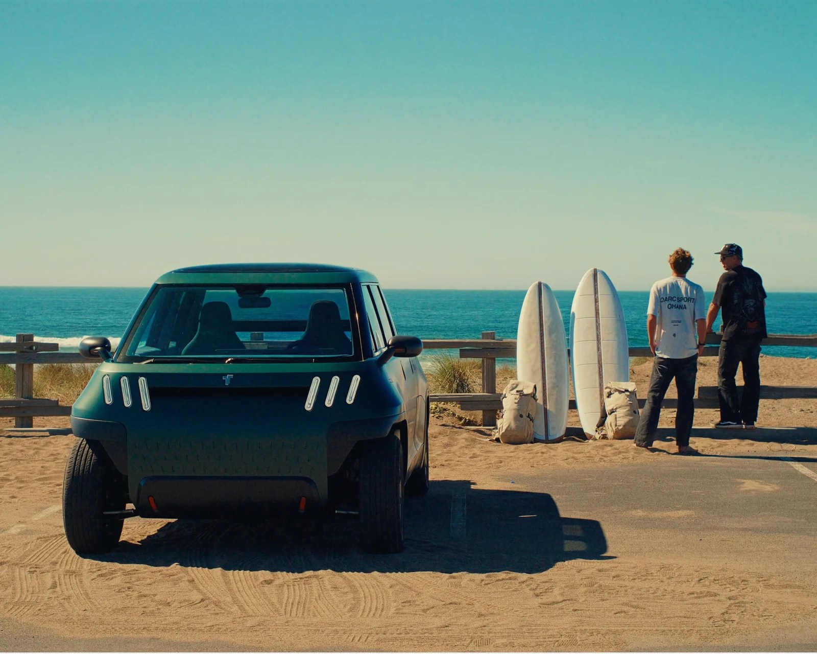 Compact dark green electric SUV parked on sandy beach near two surfboards and two people facing the ocean.