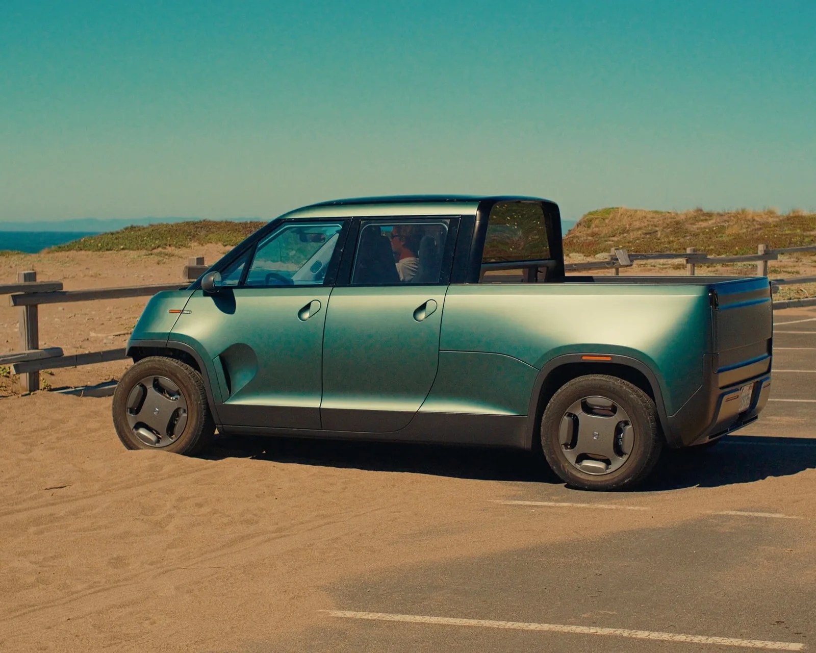 Matte green electric pickup truck with black trim parked on sandy ground near a wooden fence.