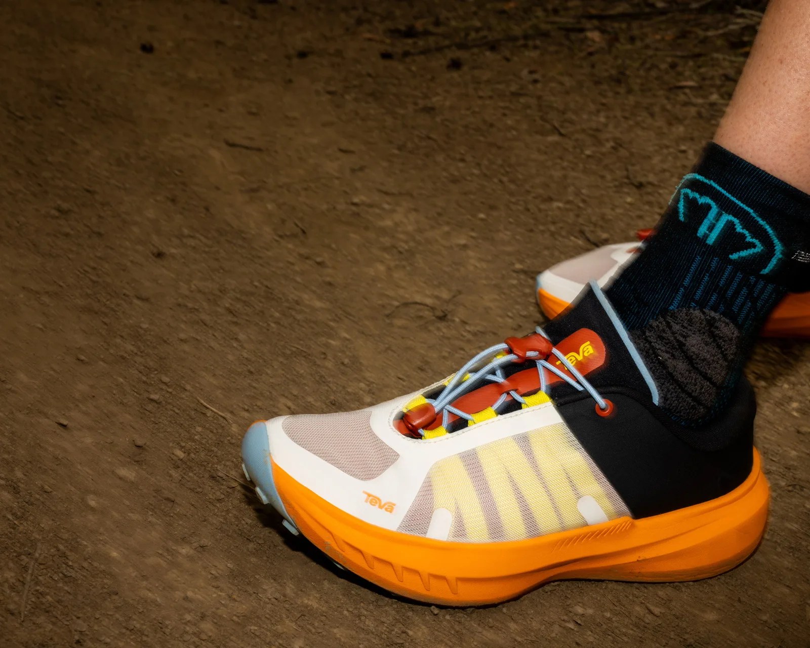 Orange, white, and yellow Teva trail running shoe worn with black and blue socks on a dirt surface.