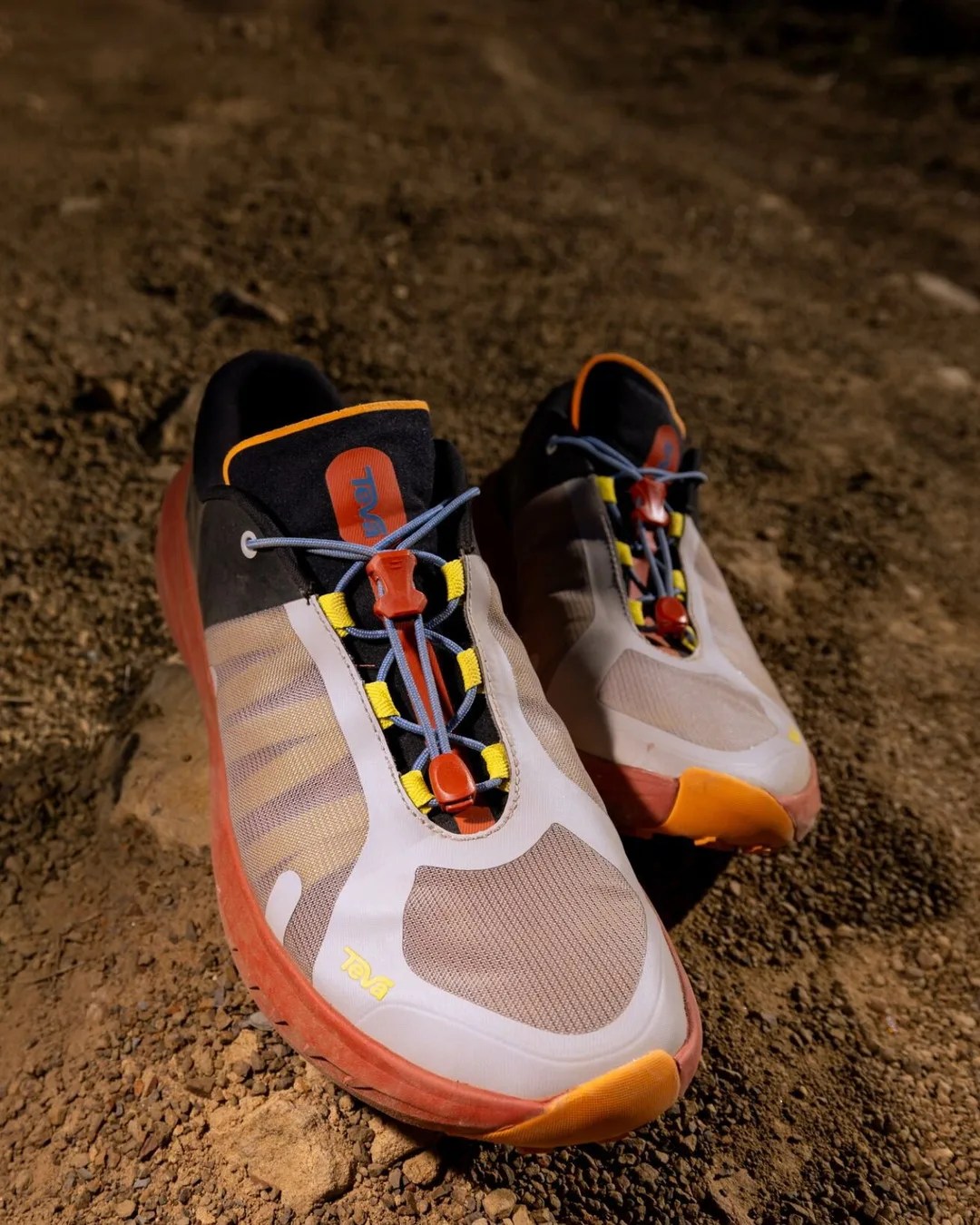 Pair of beige and orange Teva athletic shoes with blue laces on dirt ground.