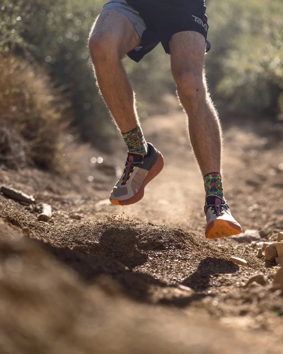 Runner's legs in colorful skull-patterned socks and gray trail running shoes jumping over a dirt path.