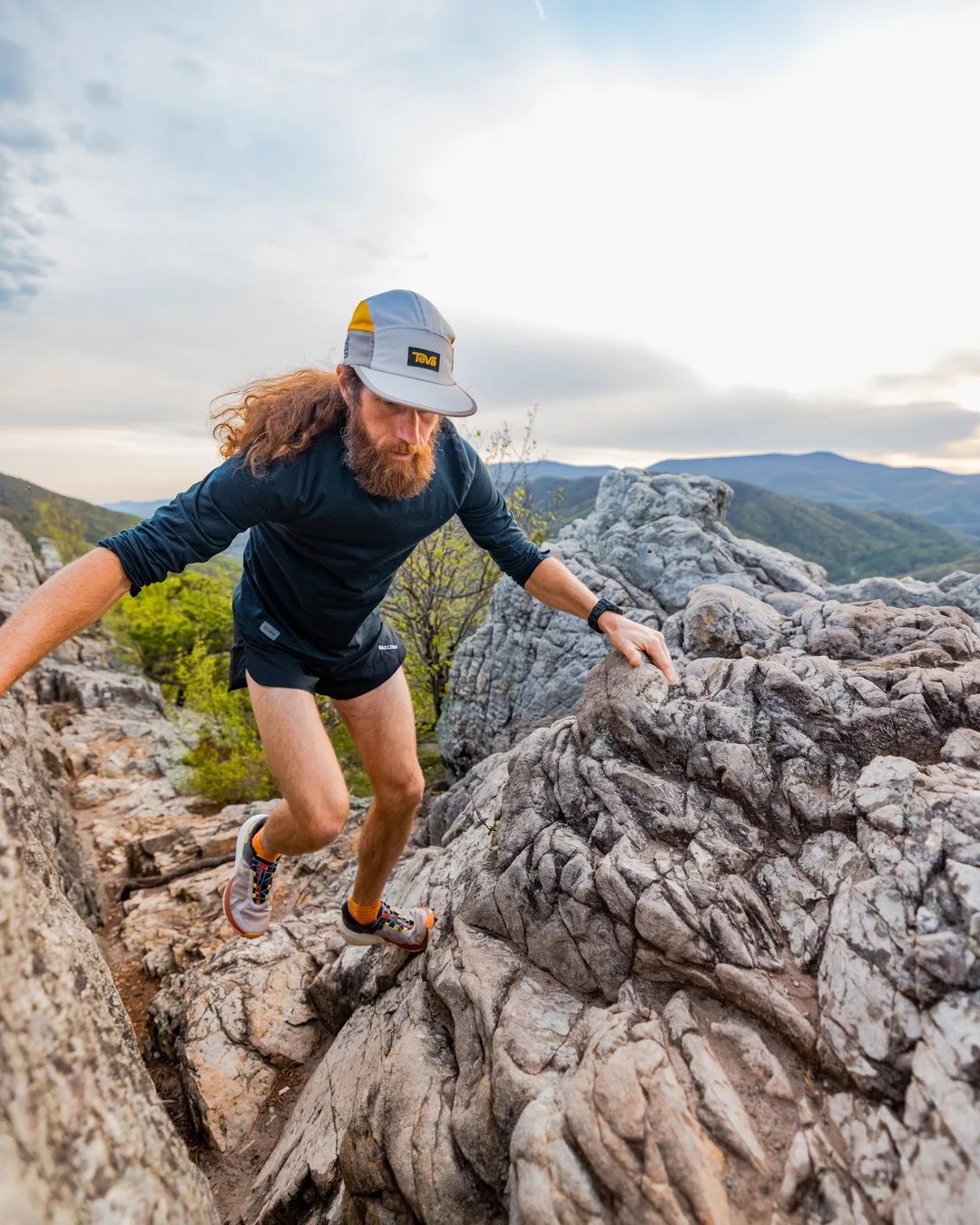 Man wearing a gray and yellow cap, black long-sleeve shirt, and shorts climbing rocky terrain outdoors.