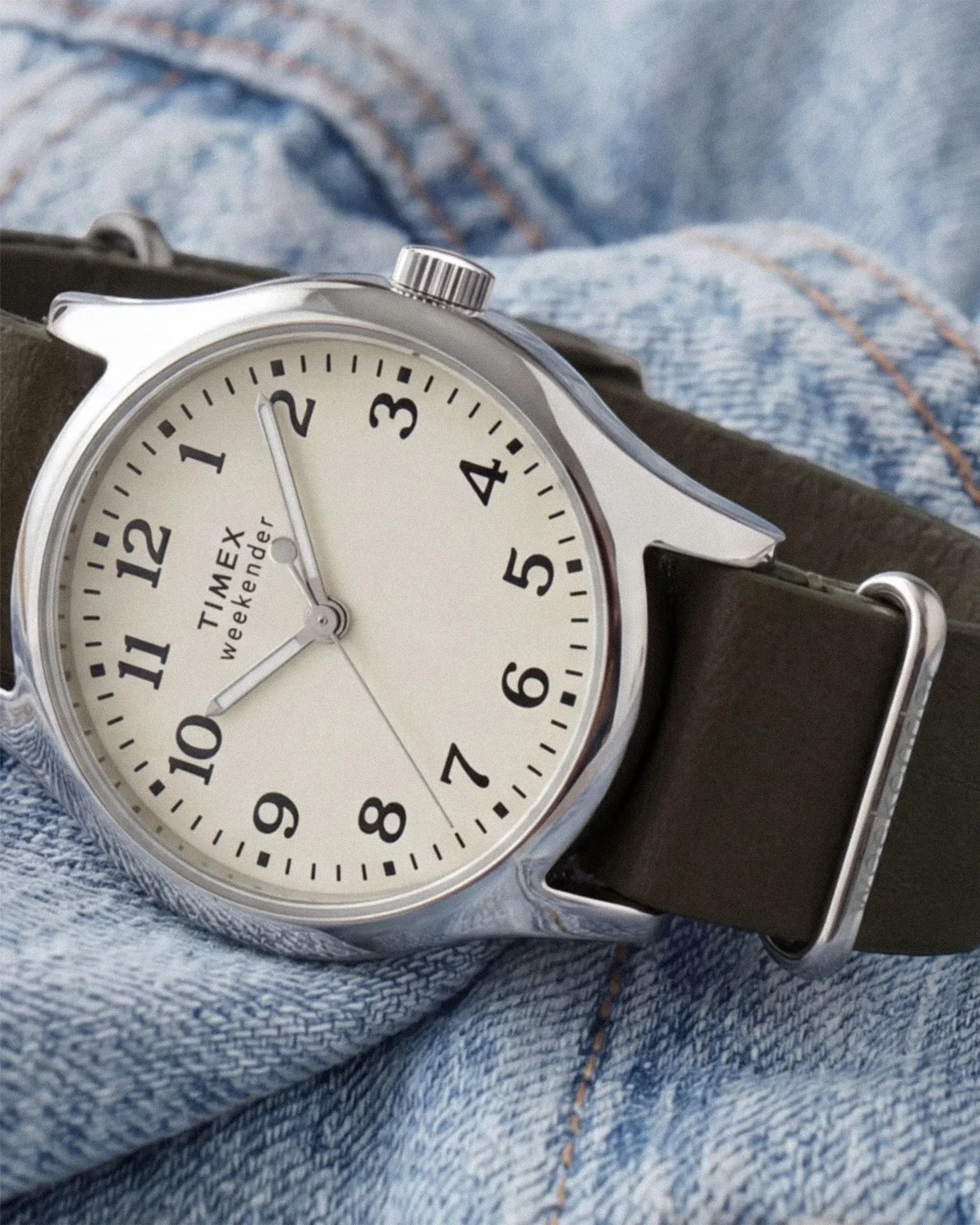 Silver Timex Weekender watch with white dial and black leather strap on blue denim fabric.