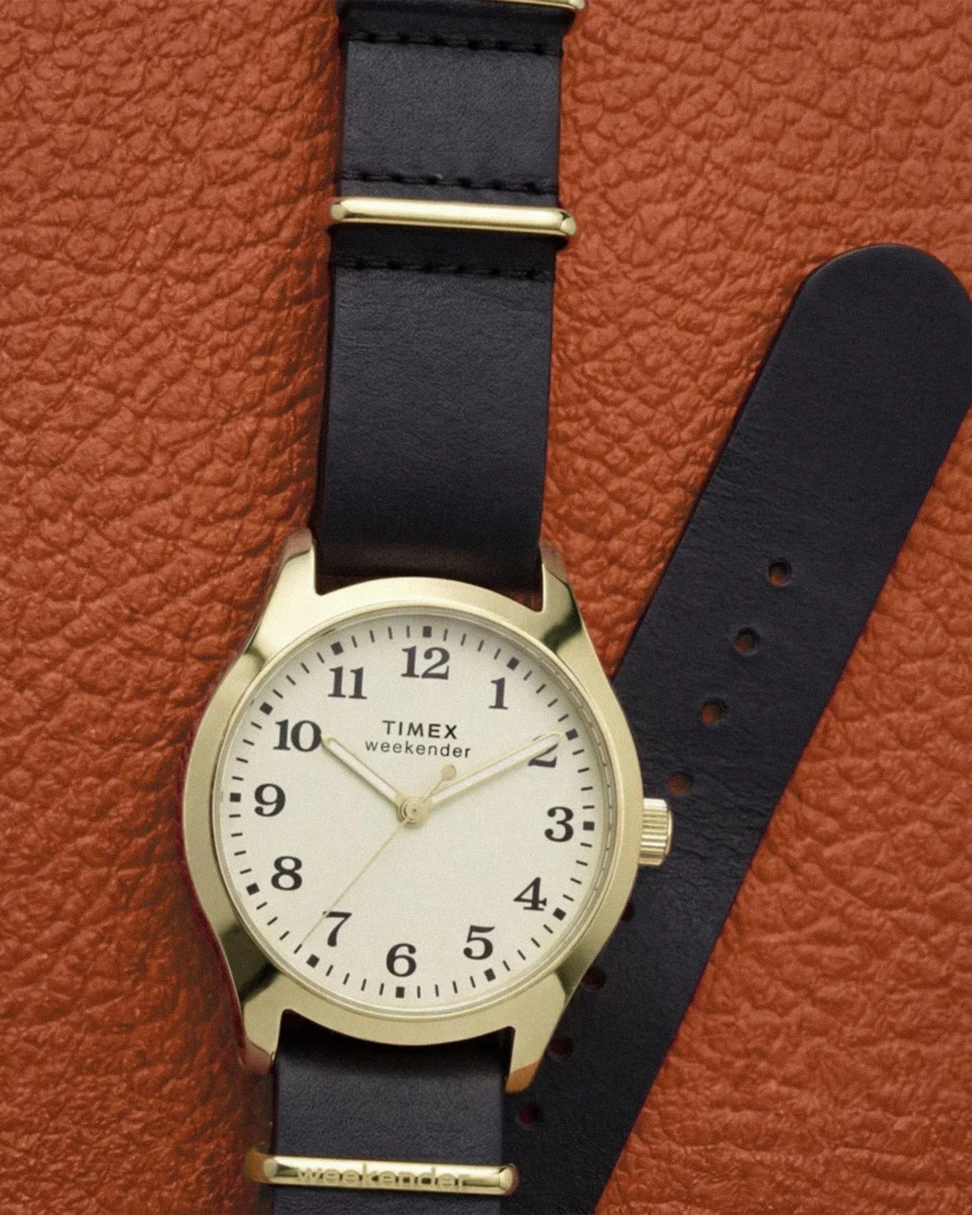 Timex Weekender watch with gold-tone case, white dial, black leather strap, and gold buckle on textured brown surface.