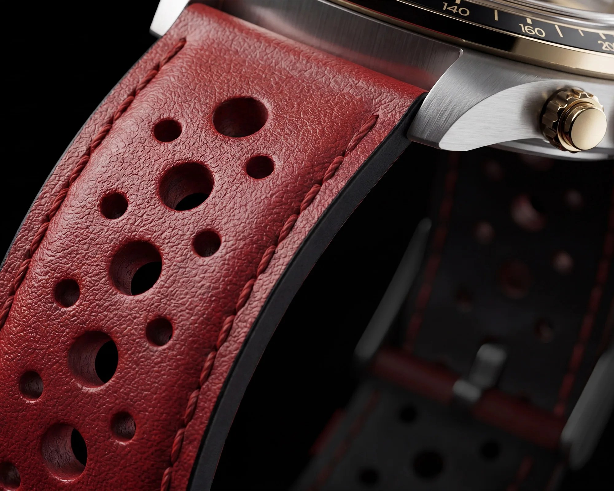 Close-up of a red perforated leather watch strap attached to a stainless steel watch case with a gold crown.
