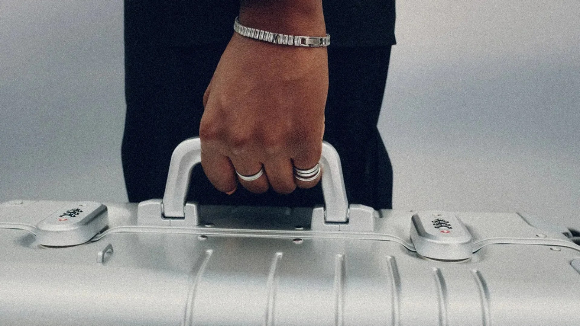 Hand wearing silver rings and a bracelet gripping the handle of a silver hard-shell suitcase.