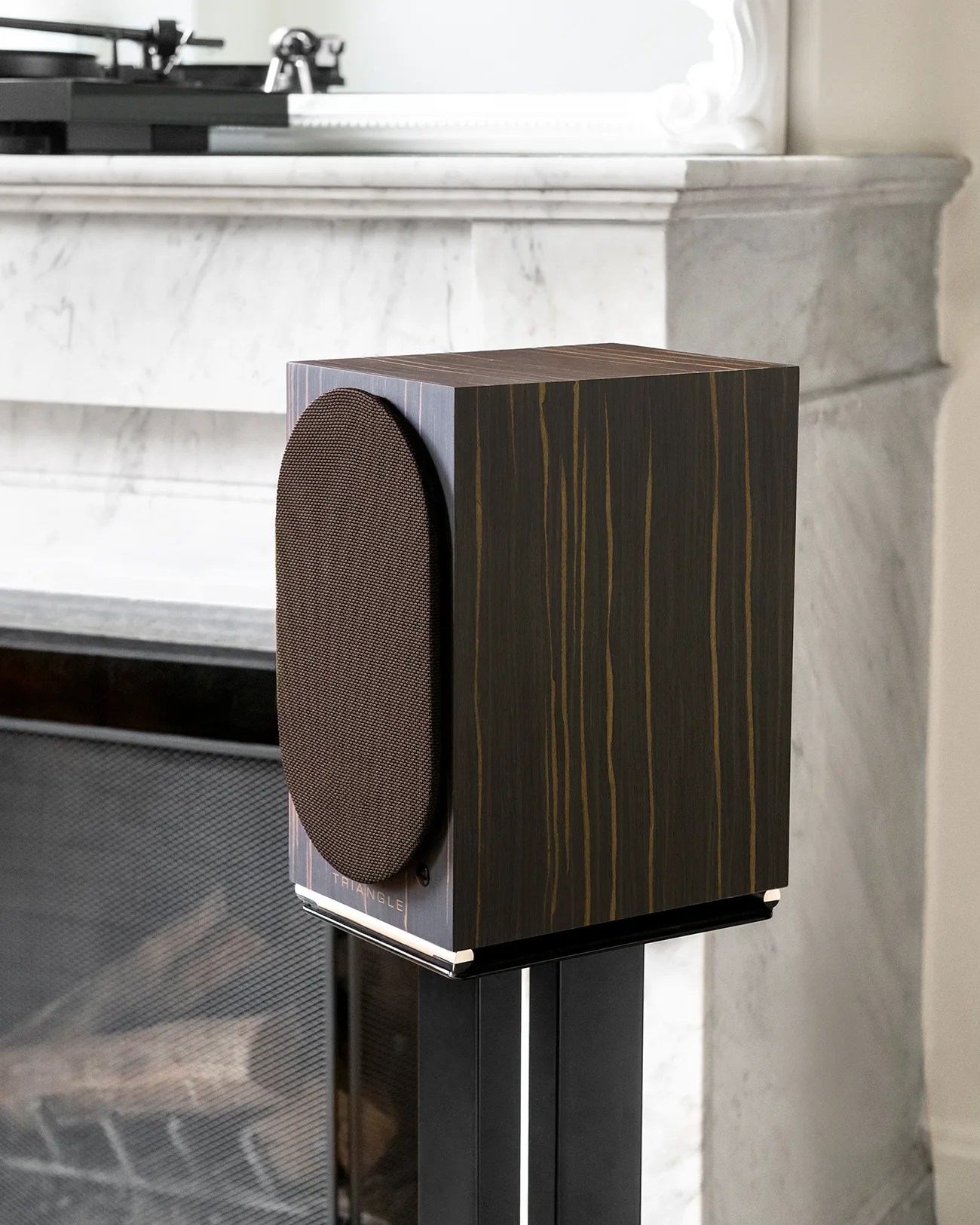 Dark wood cube speaker with a brown fabric grille on a black metal stand in front of a marble fireplace.