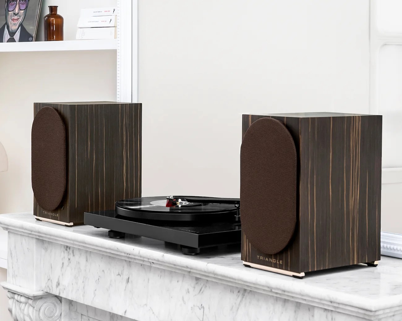 Pair of dark wood bookshelf speakers with brown fabric grilles flanking a black turntable on a white marble mantel.