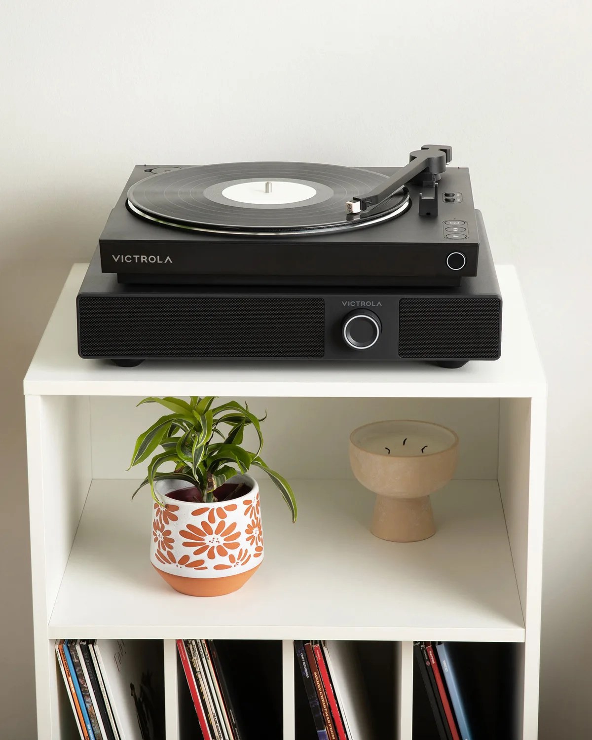 Black Victrola turntable with vinyl record on top of matching speaker on white shelf with plant and candle below.
