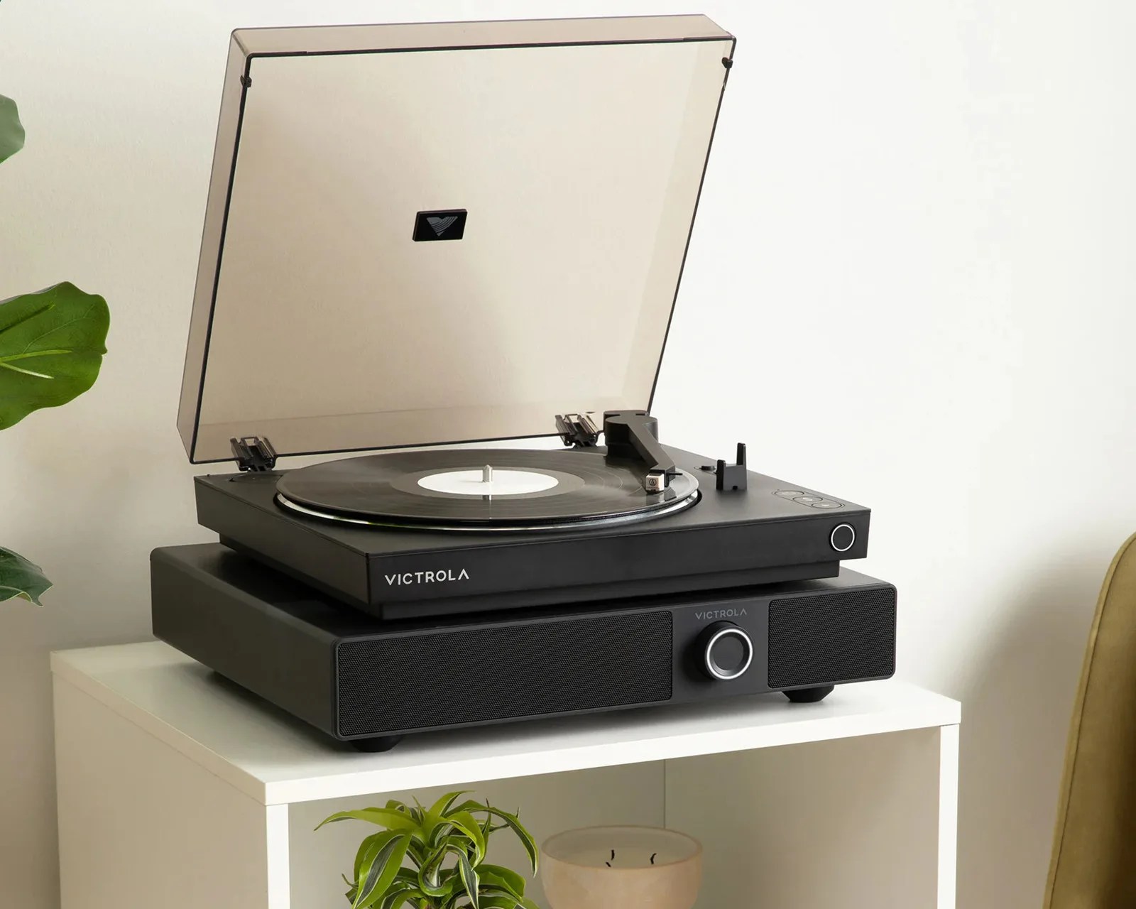 Black Victrola turntable with transparent dust cover open, playing a vinyl record on a white table.