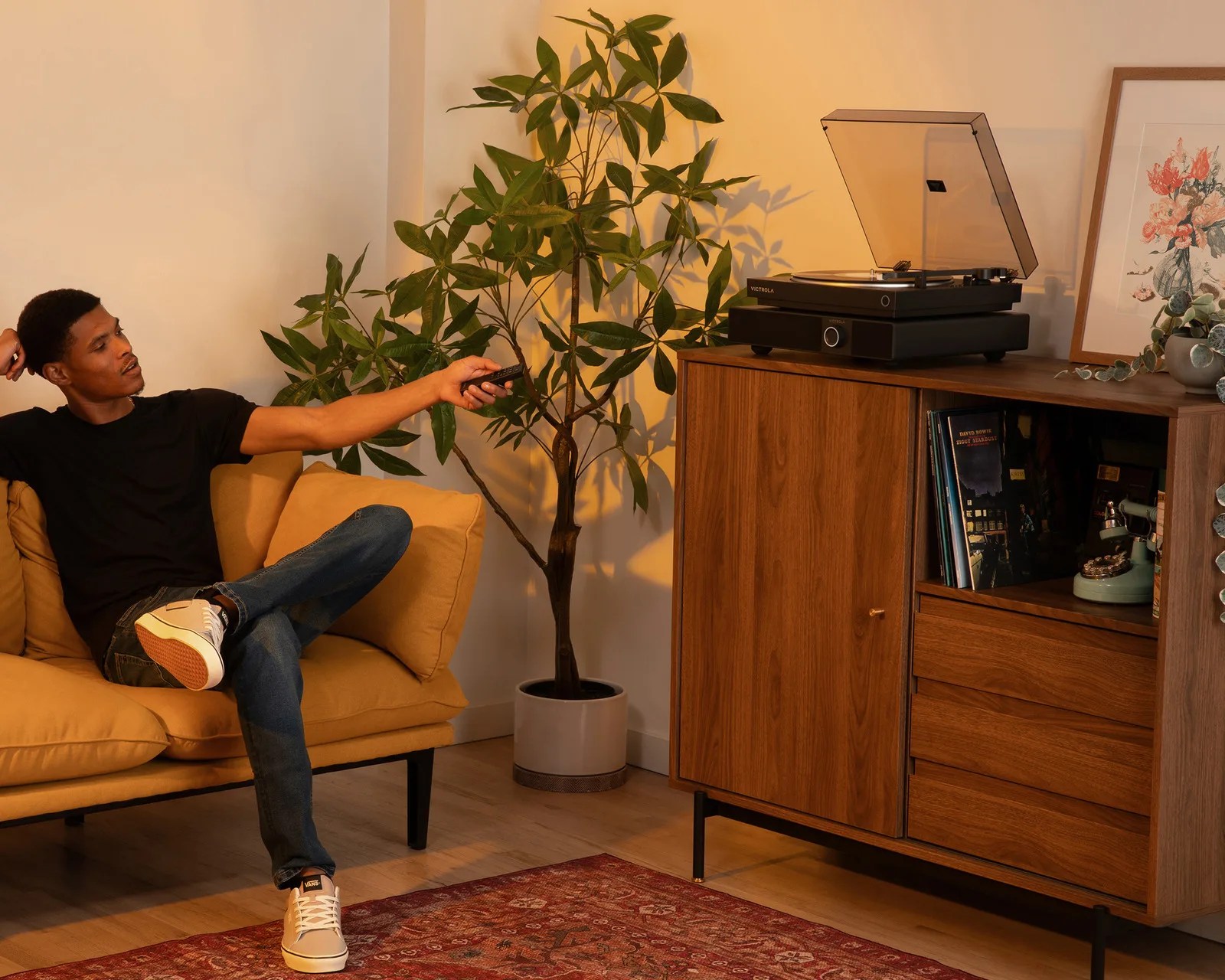 Man in black t-shirt and jeans sitting on mustard sofa holding remote, next to a potted plant and wooden cabinet with a turntable.