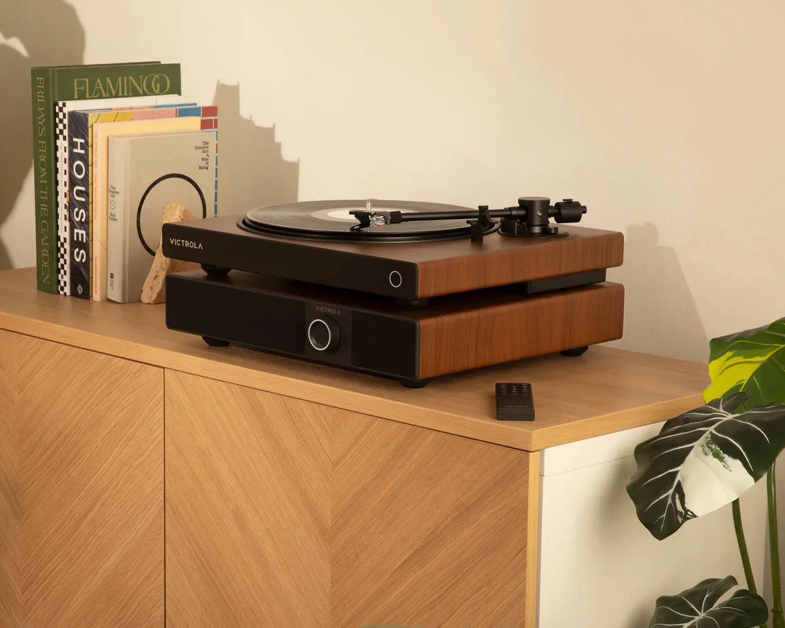 Wood-finished Victrola turntable with black tonearm and speaker on a wooden cabinet with books and a remote.