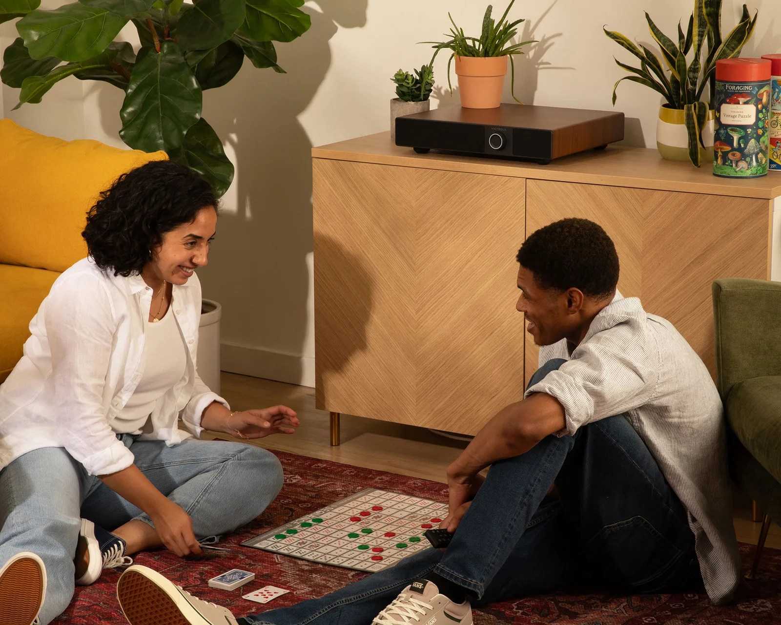 Two people sitting on a red patterned rug playing a board game with red and green pieces.