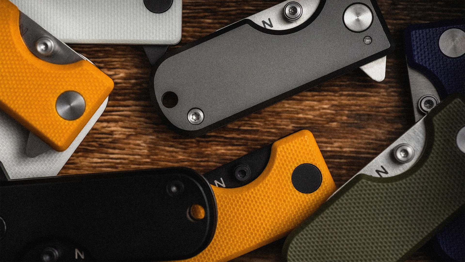 Close-up of various folding knives with textured handles in yellow, black, white, gray, blue, and green on a wooden surface.