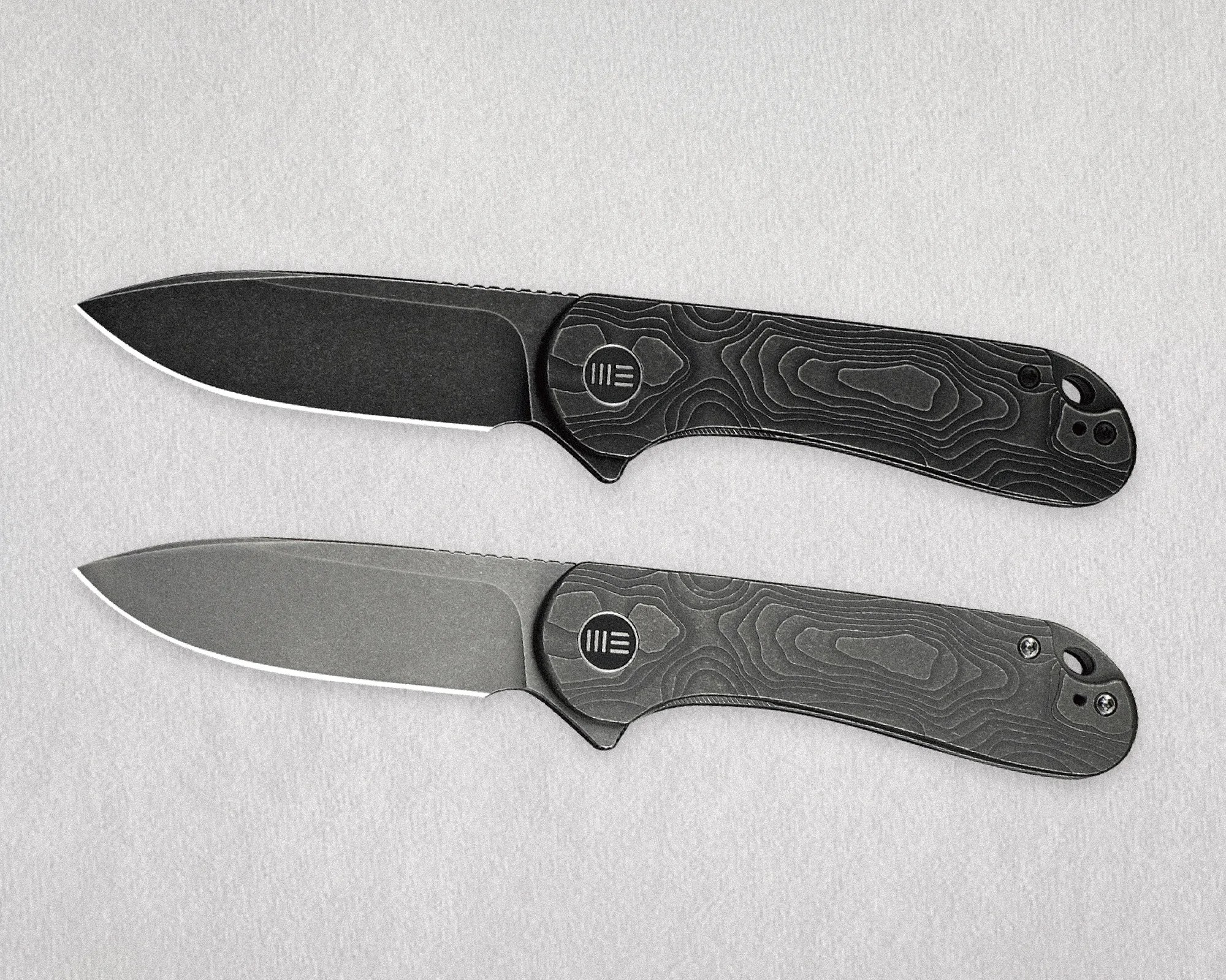 Two black folding knives with topographic pattern handles and partially serrated blades on a light gray surface.