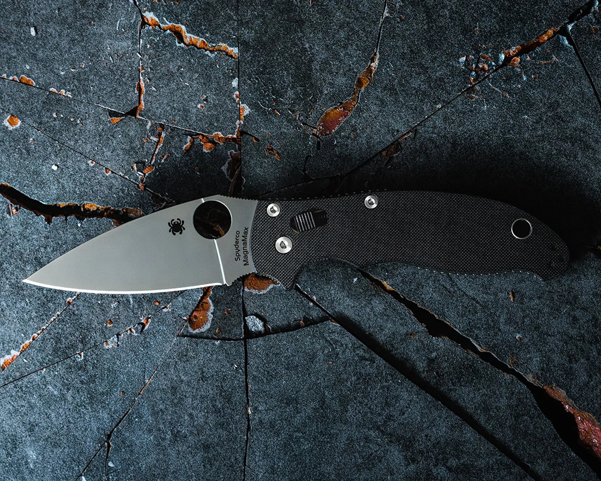 Spyderco MagnaMax folding knife with a silver blade and black textured handle on cracked dark stone surface.