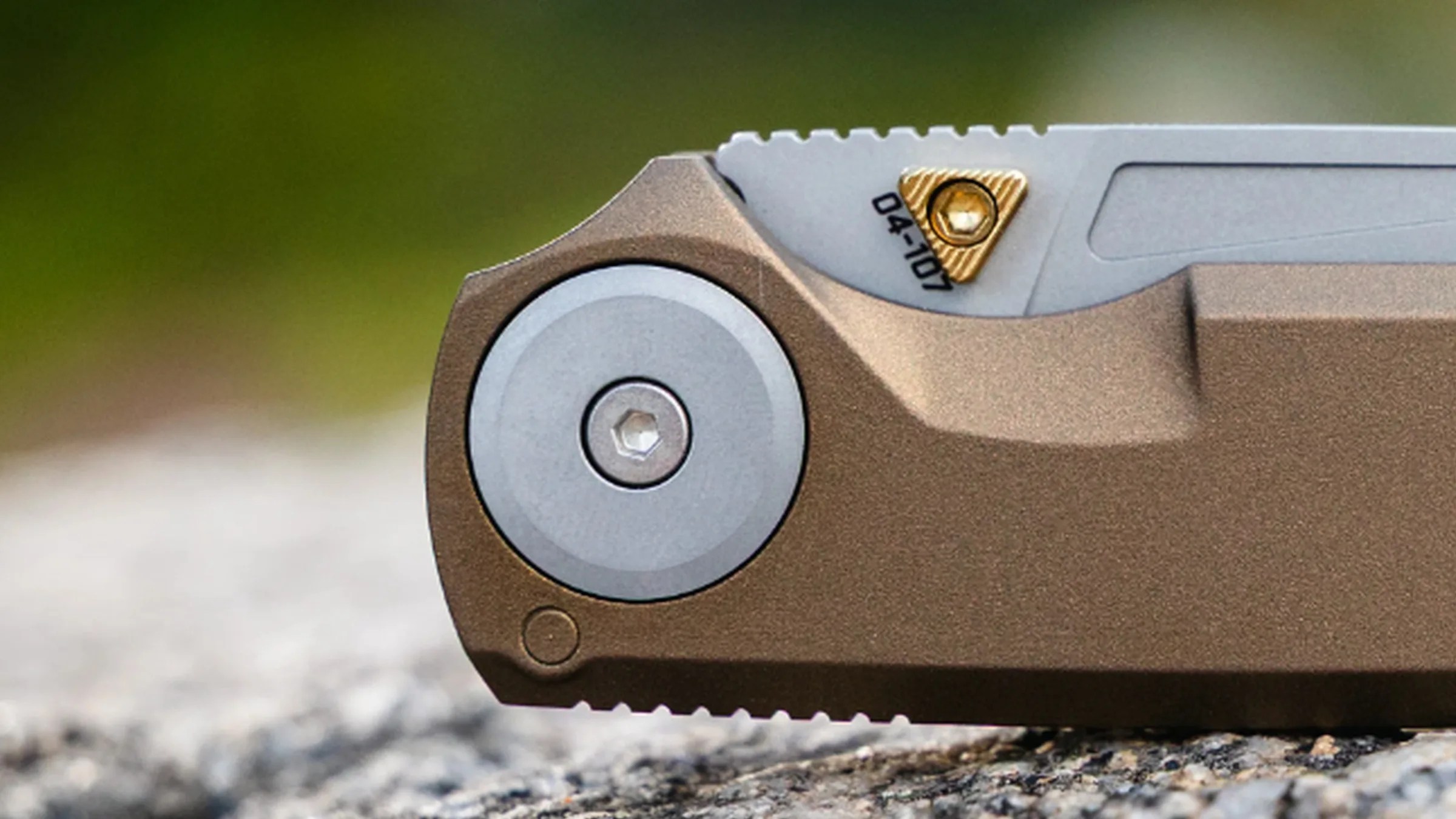 Close-up of a folding knife pivot with bronze handle and silver blade featuring a gold triangular screw.
