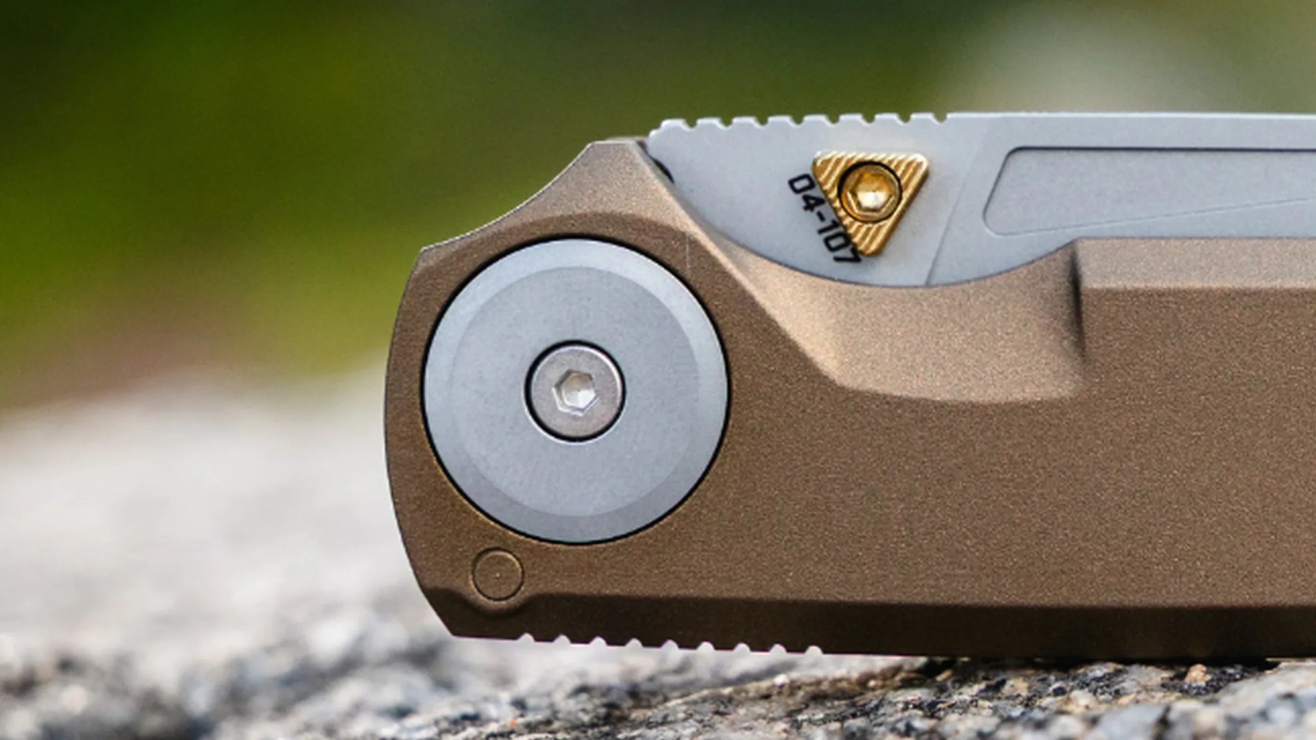 Close-up of a folding knife pivot with bronze handle and silver blade featuring a gold triangular screw.