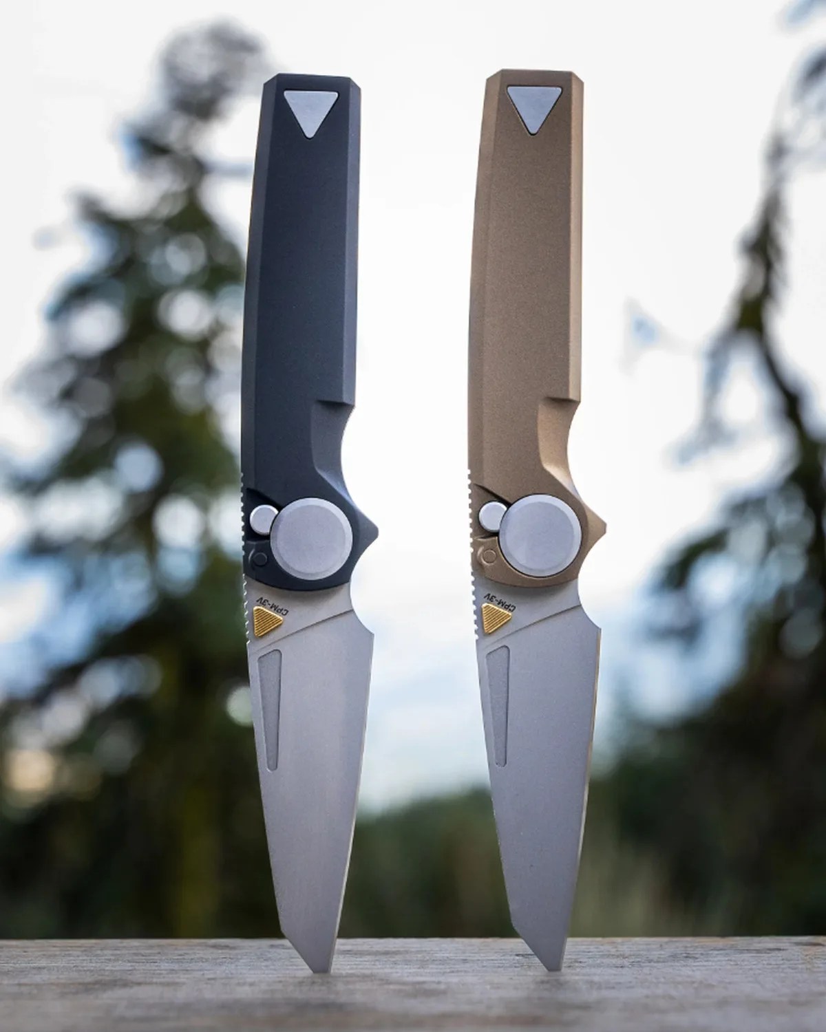 Two folding knives with silver blades and triangular metal inlays on the handles, one black and one tan, standing upright on a wooden surface.