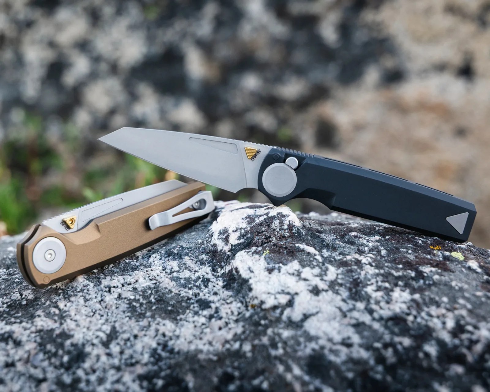 Two folding knives with silver blades, one with a black handle and the other with a bronze handle, resting on a rock.