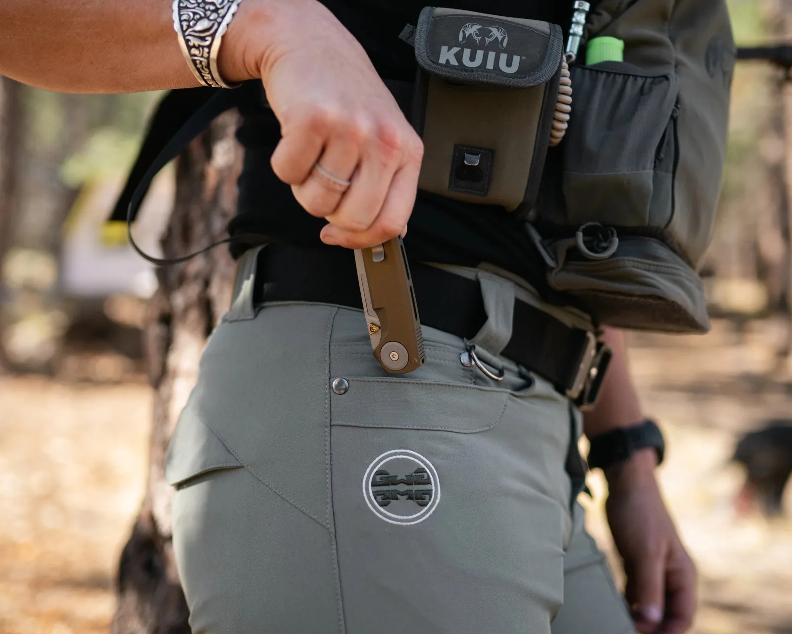 Person wearing gray pants with a circular logo, holding a small folding knife near the pocket, and carrying a green Kuiu pouch.