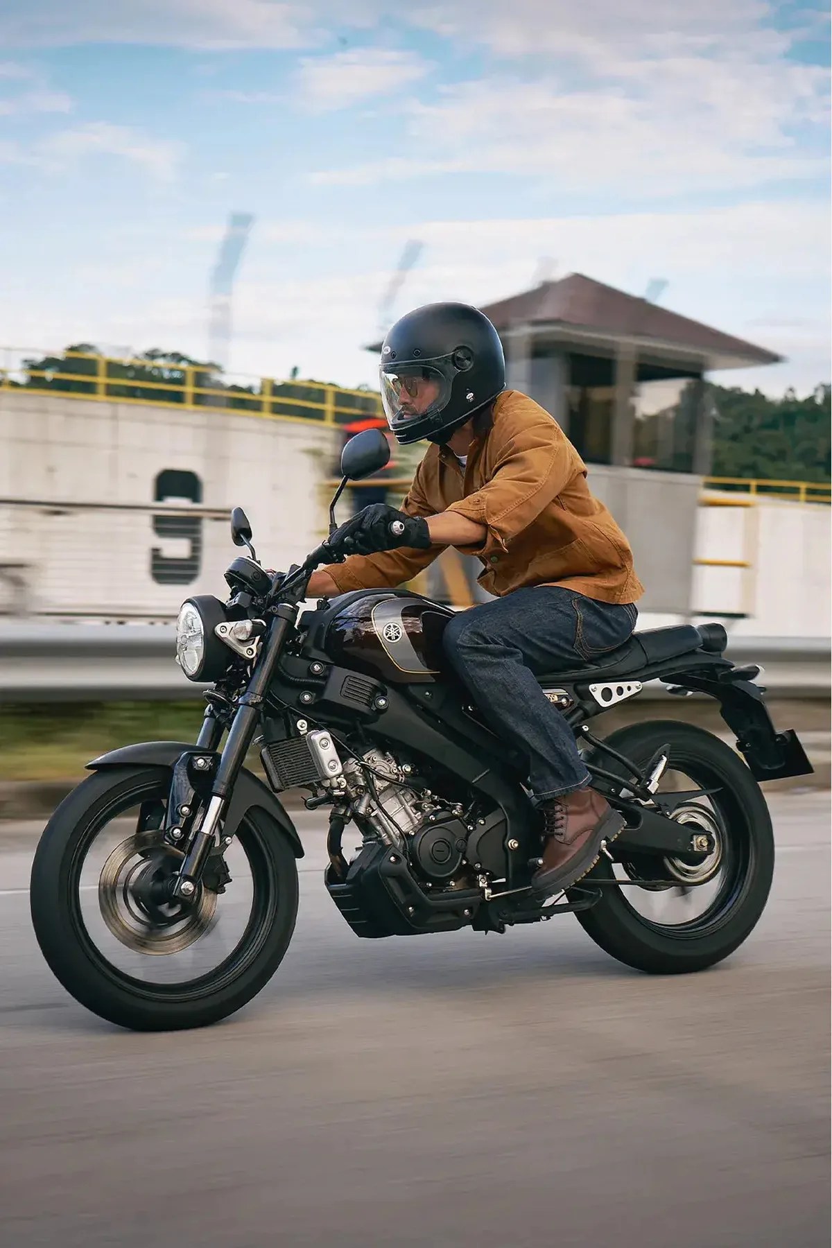 Person wearing a black helmet and brown jacket riding a black Yamaha motorcycle on a road.