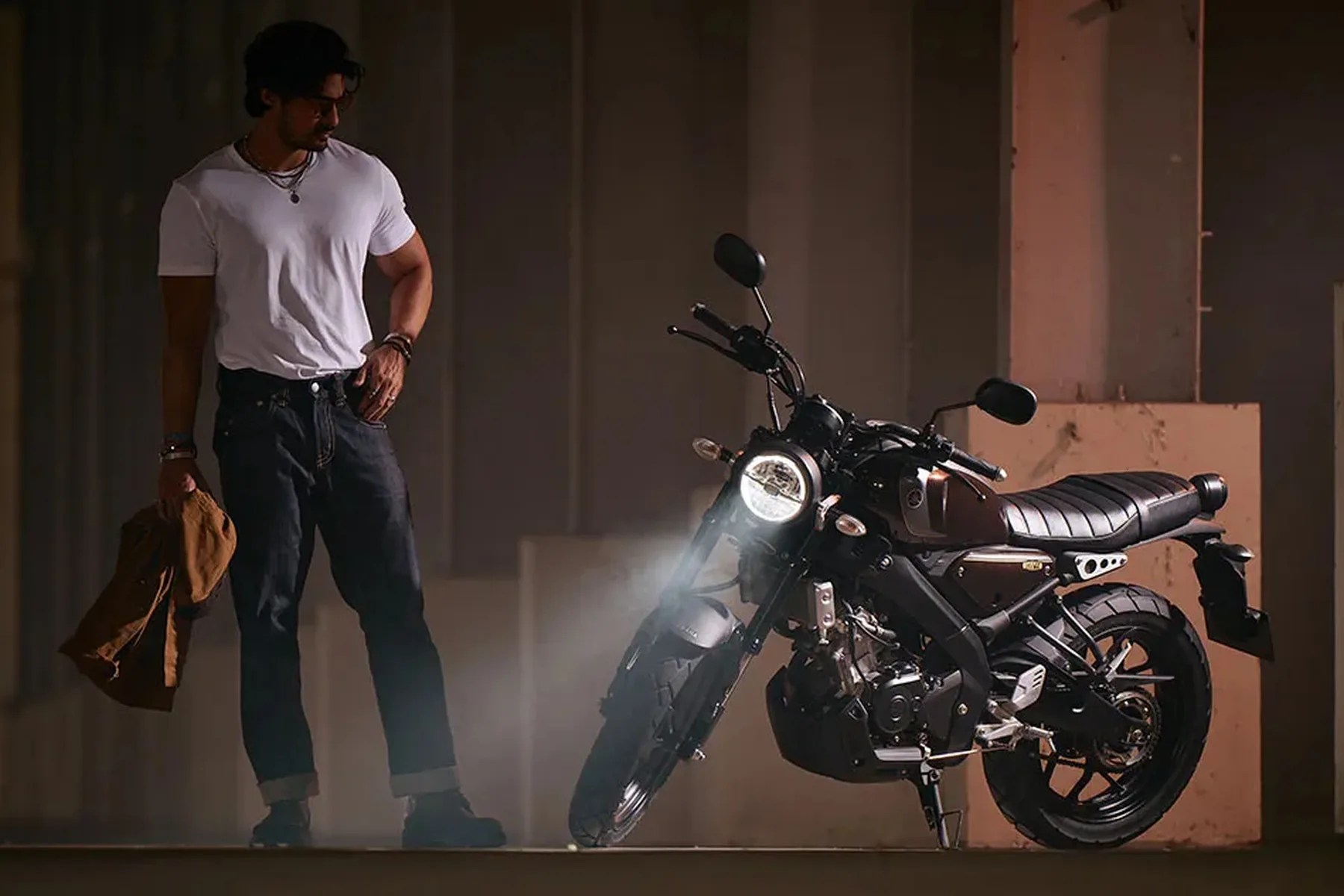 Man in white t-shirt and dark jeans holding a brown jacket standing next to a black motorcycle with its headlight on.