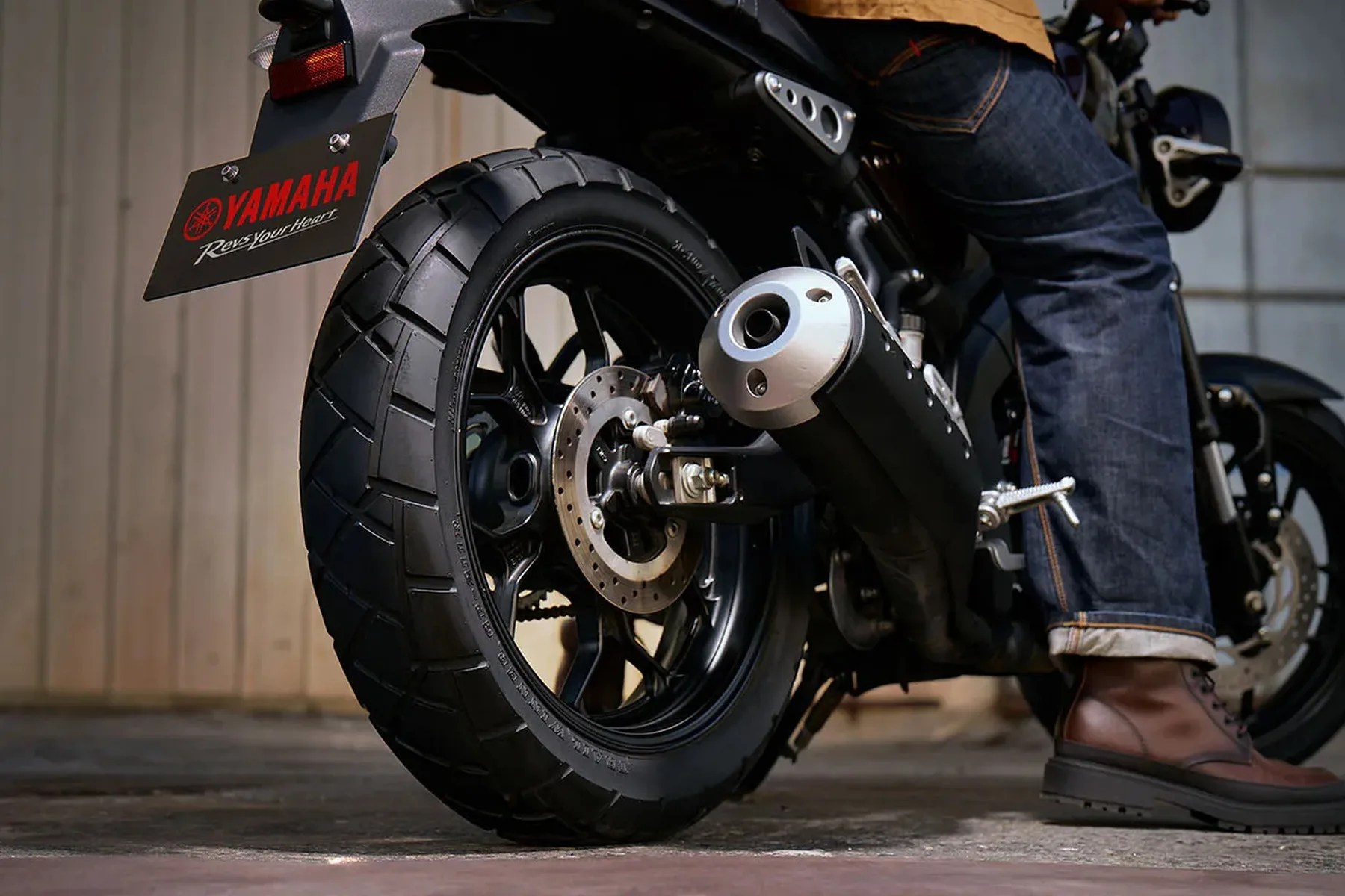 Rear view of a Yamaha motorcycle showing the rear tire, exhaust pipe, and a person wearing jeans and brown boots sitting on it.
