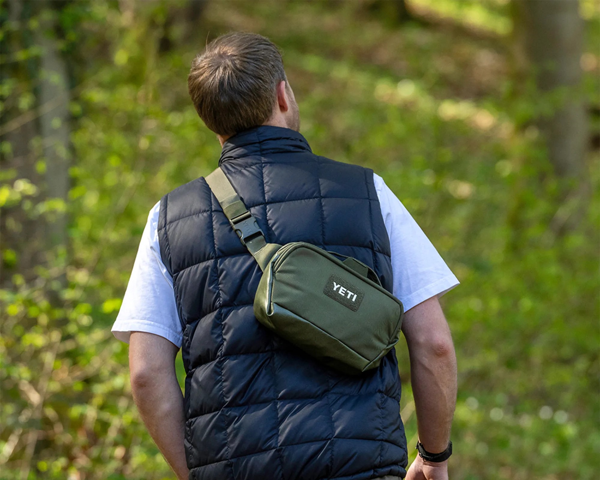 Man wearing a navy blue quilted vest and white t-shirt with an olive green YETI crossbody bag outdoors.