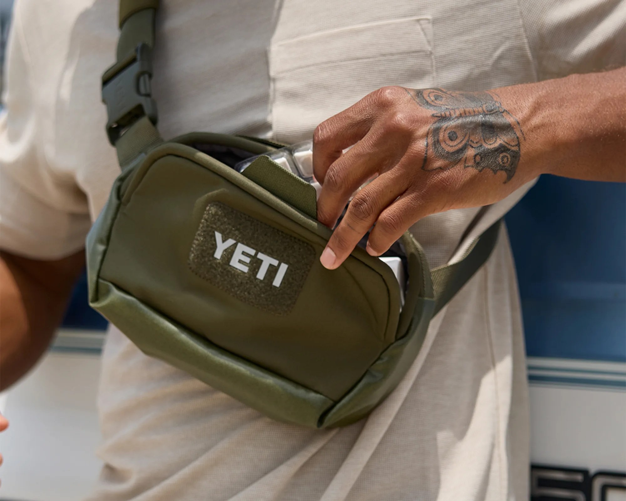 Olive green YETI crossbody bag worn over a beige shirt with a hand featuring a butterfly tattoo reaching into the bag.
