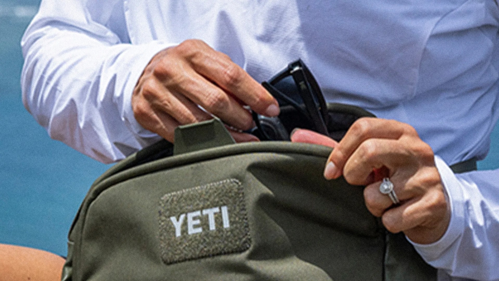 Hands placing black sunglasses into an olive green YETI bag worn by a person in a white long-sleeve shirt.
