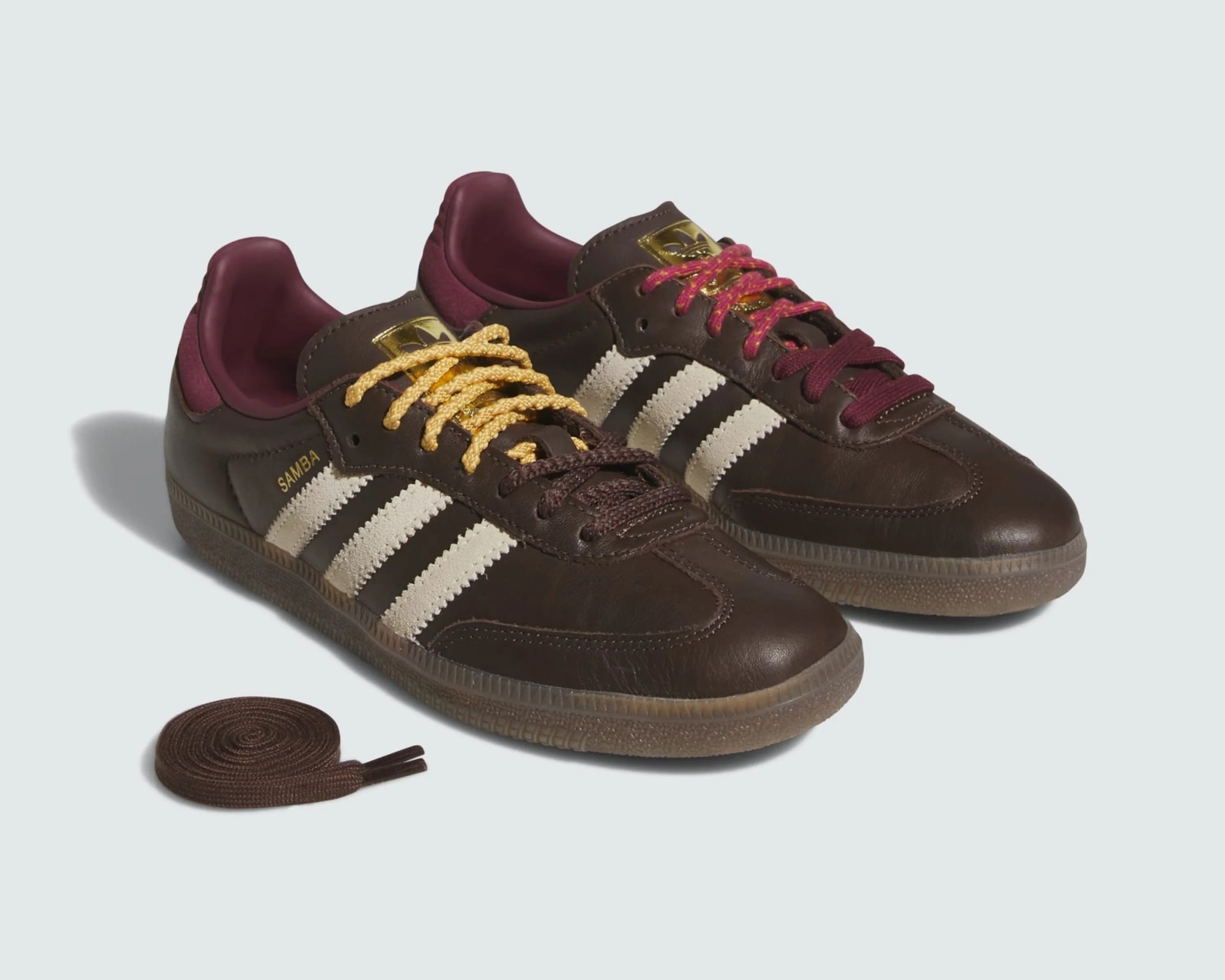 Pair of brown Adidas Samba sneakers with white stripes, gold logo, and mismatched yellow and red laces, with extra brown laces nearby.