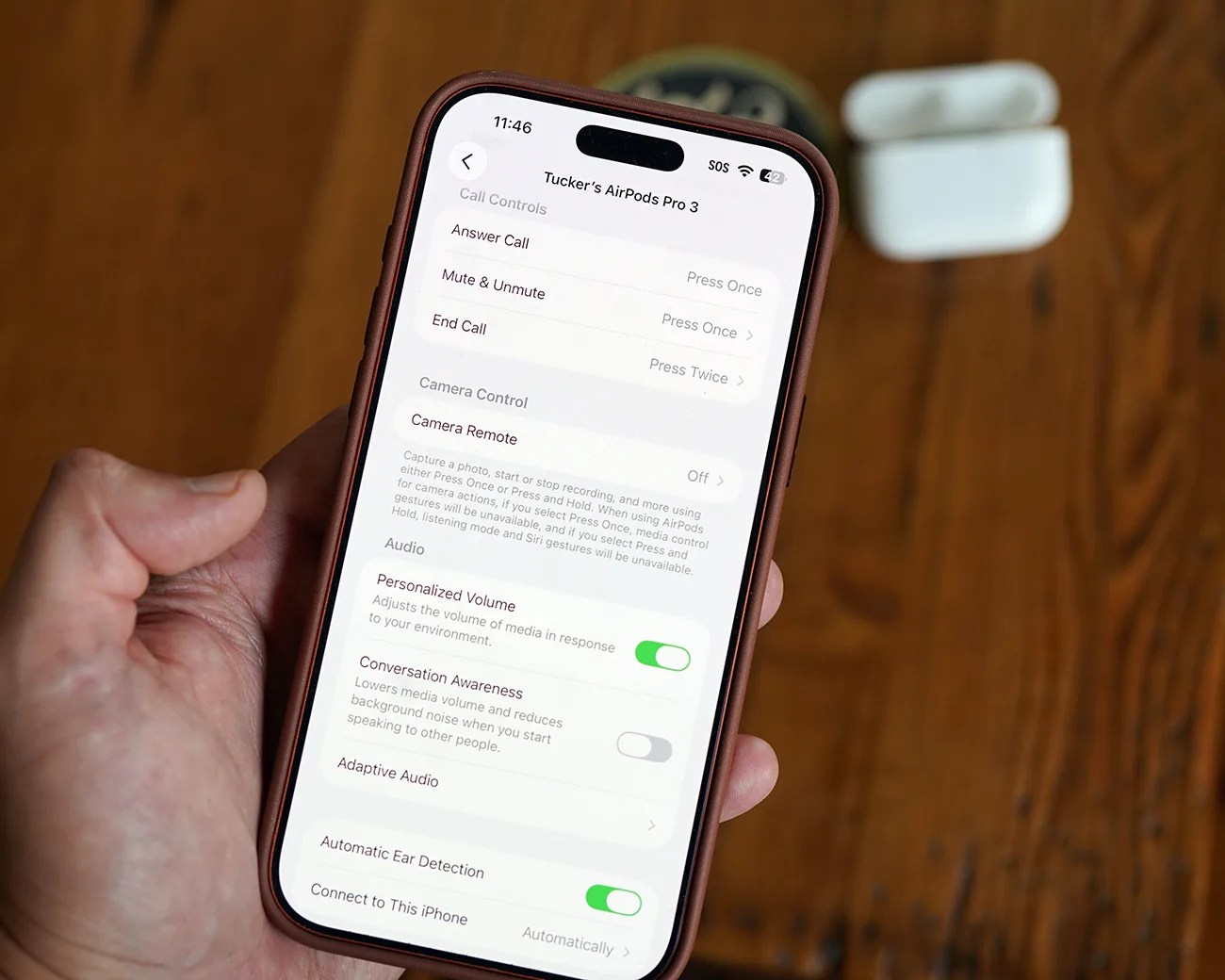 Hand holding a smartphone displaying AirPods Pro 3 settings with a white AirPods case on a wooden surface.
