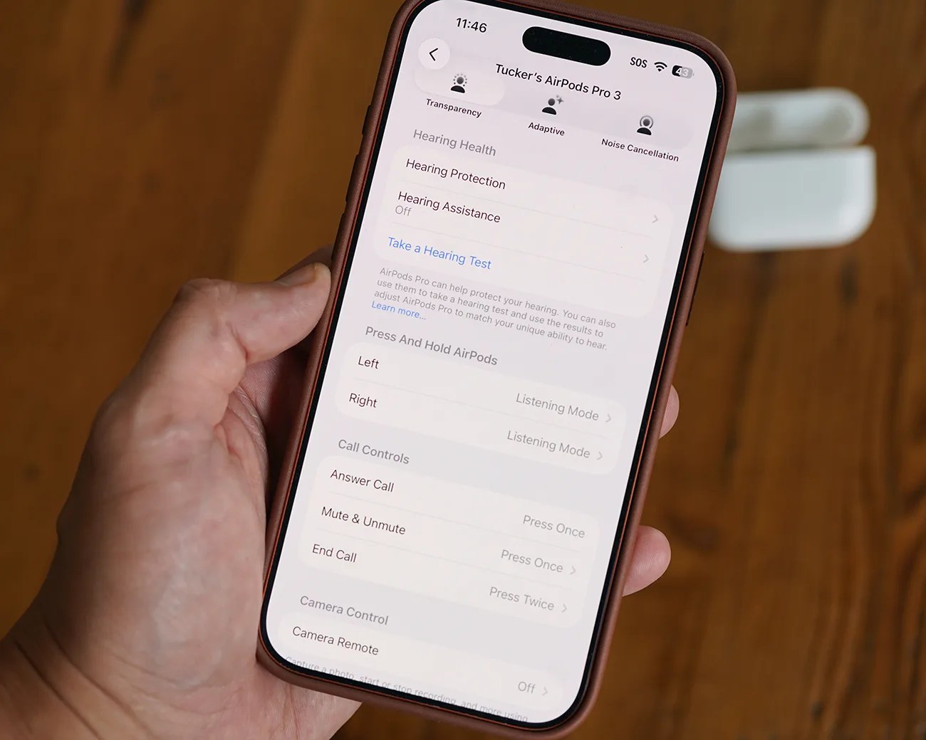 Smartphone screen showing AirPods Pro 3 settings with hearing health and call controls, held in a hand over a wooden surface.